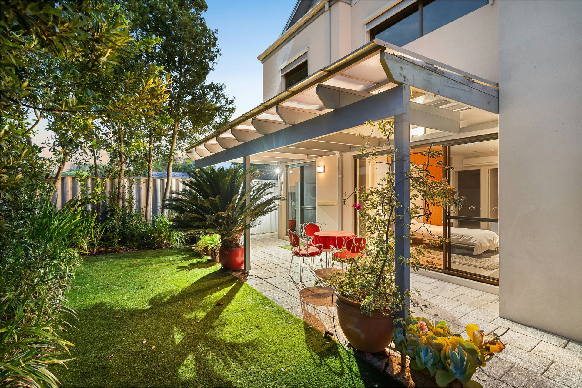 Backyard patio with glass canopy, red chairs, and lush green lawn beside a modern house