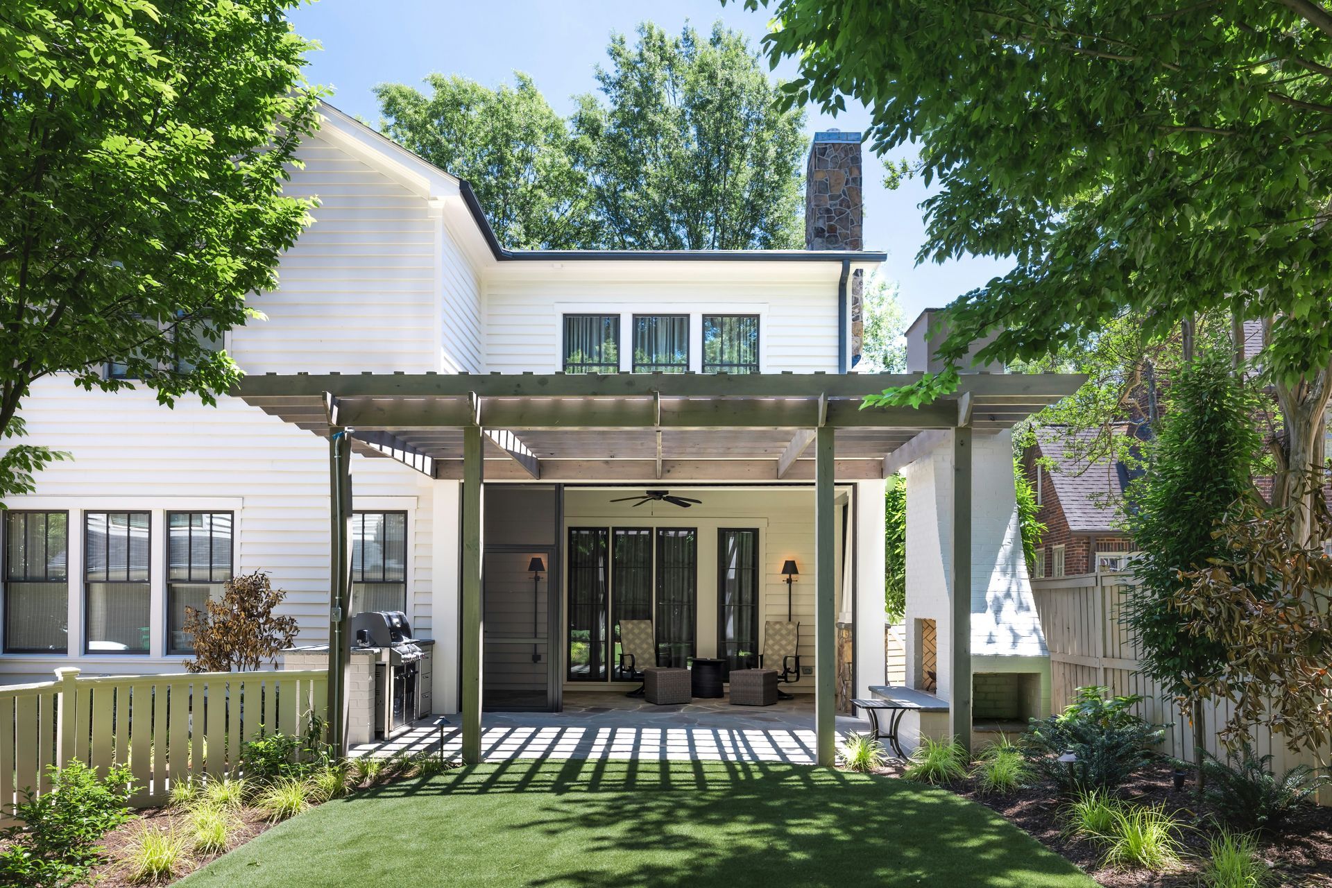 Sunlit backyard patio with pergola, white house exterior, and green lawn shaded by trees