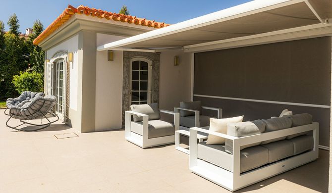 Patio with white sectional sofa, cushioned chairs, and a closed gray awning beside a stucco house