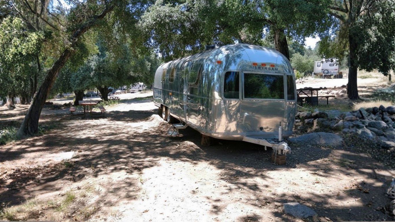 Tan RV with slide-out extended in a gravel campsite. Blue sky, palm trees.