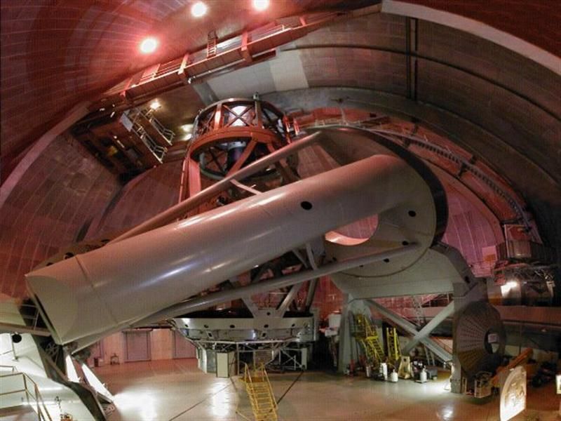 Inside a dome, a large telescope is mounted, its white tube angled. Red lighting illuminates the interior.