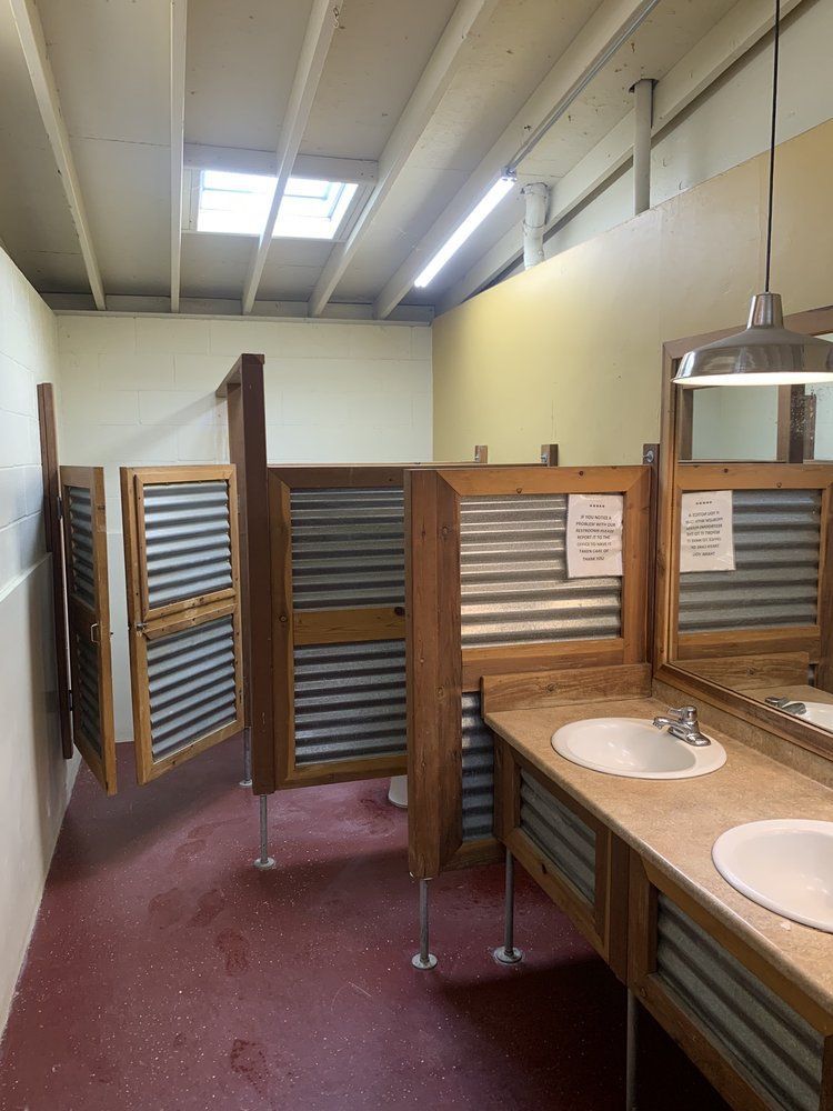 Public restroom with wood and corrugated metal stalls and sinks, red floor, and overhead lighting.