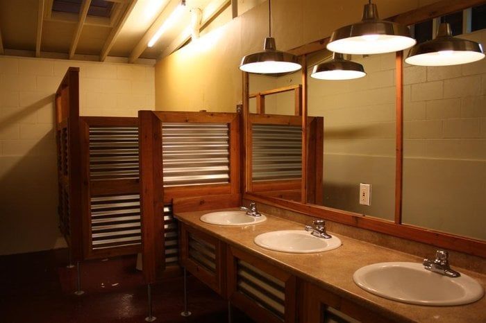 Rustic bathroom with three sinks, metal-accented wooden stalls, and hanging lights.