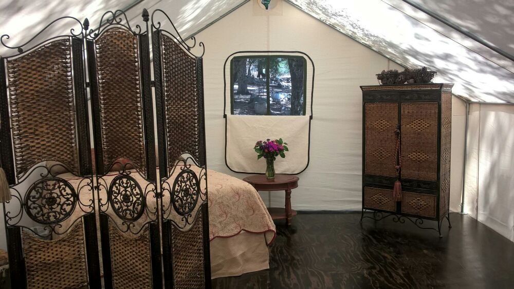 Interior of a tent with bed, folding screen, wardrobe, and table with flowers. White walls and a dark wooden floor.