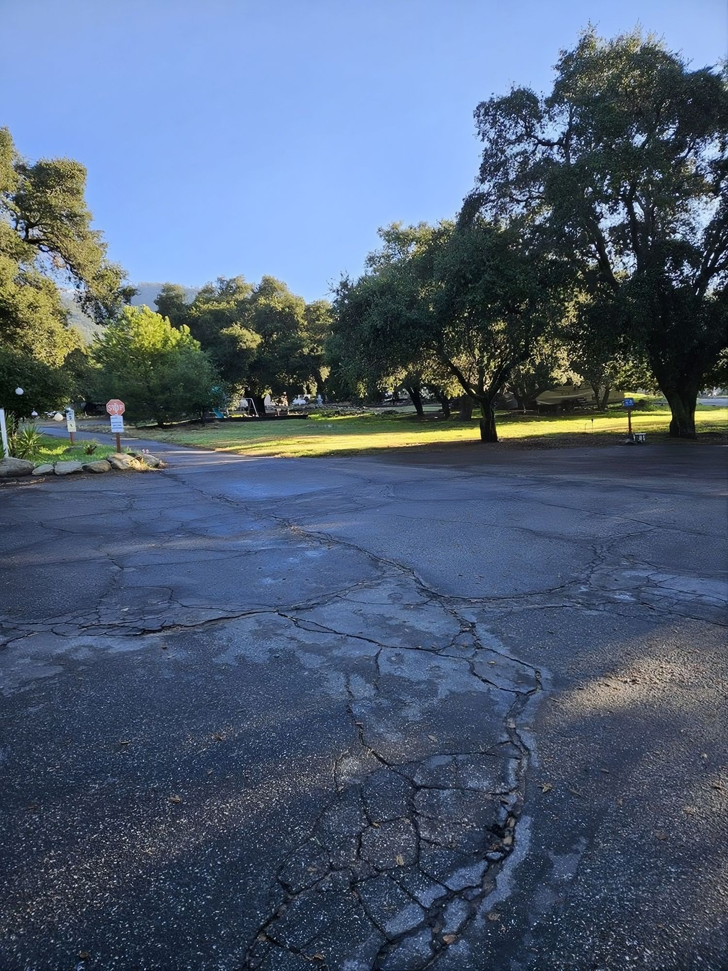 Cracked asphalt road leads to a green space with trees, mountains in the distance, and a blue sky.