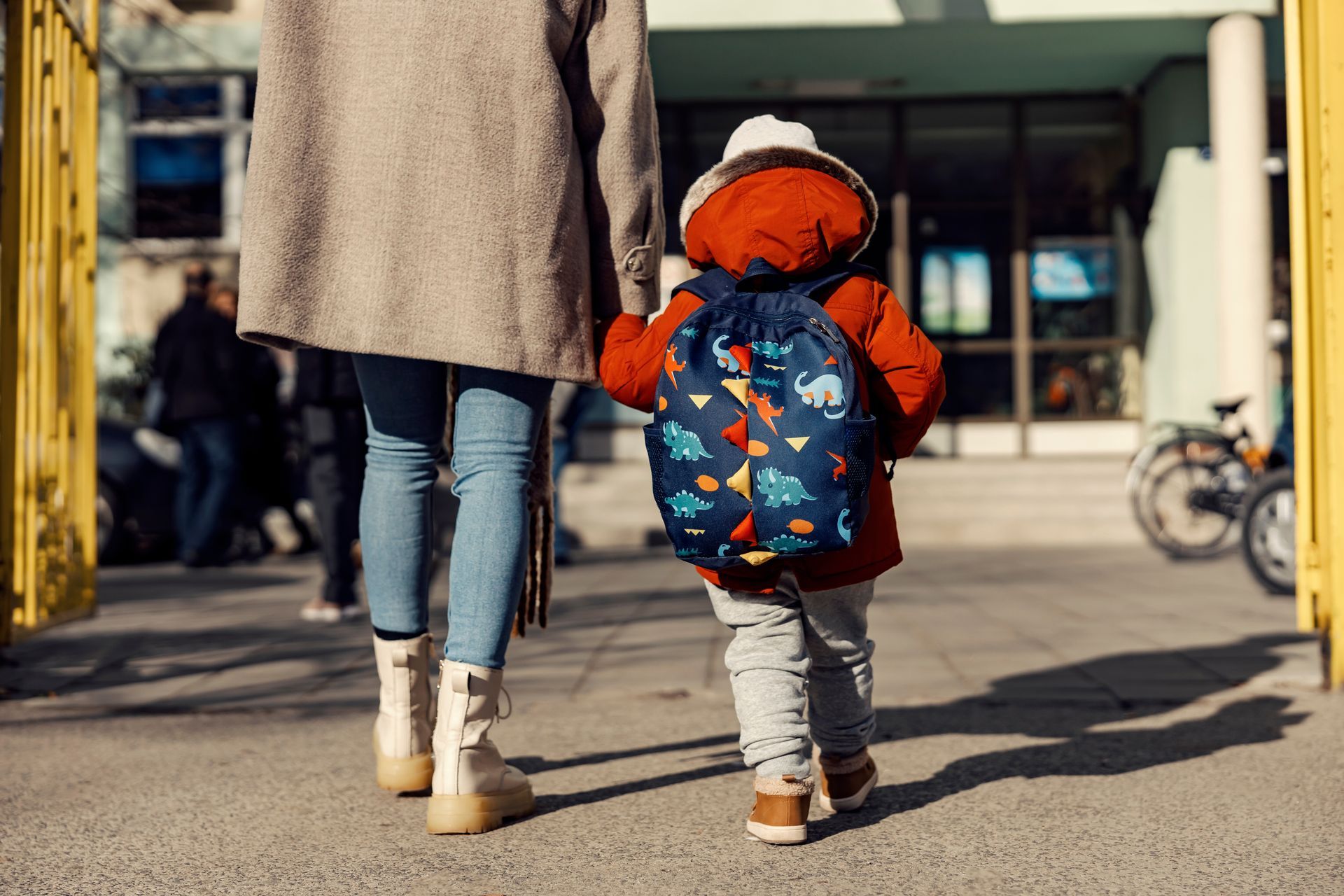 Adult and child walking toward a building, child wearing an orange jacket and blue backpack