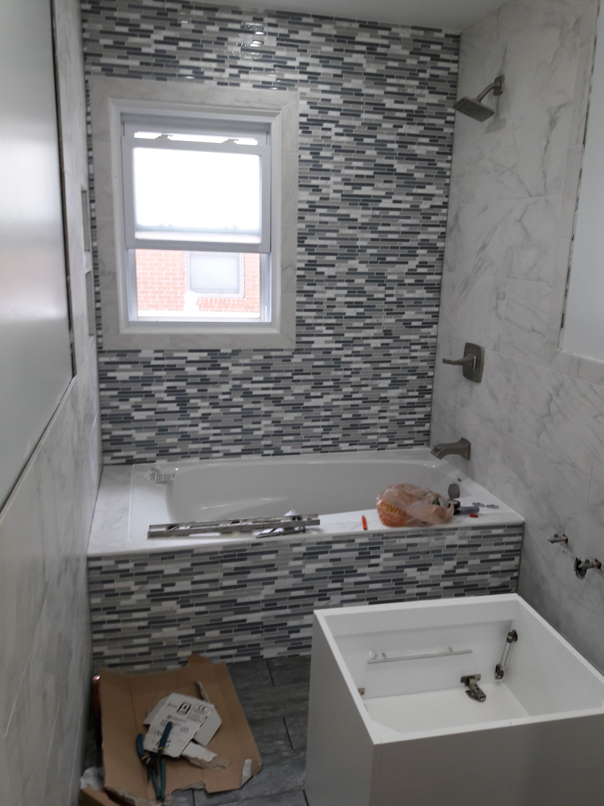 A bathroom under renovation featuring a bathtub with gray mosaic tiled surround and wall, and a white vanity cabinet.