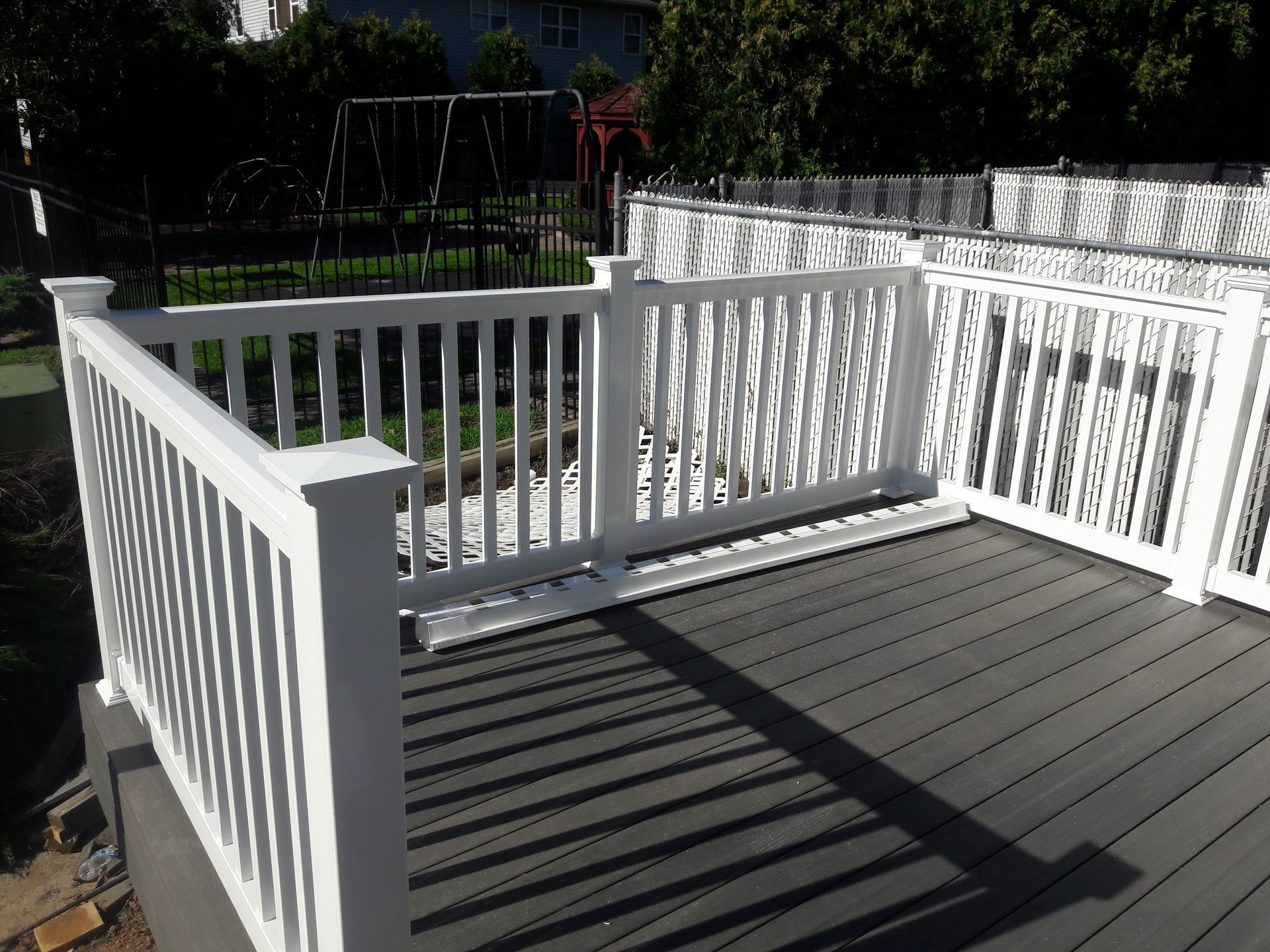 A white vinyl railing encloses a corner of a grey composite deck, set against a background of trees and a chain-link fence.
