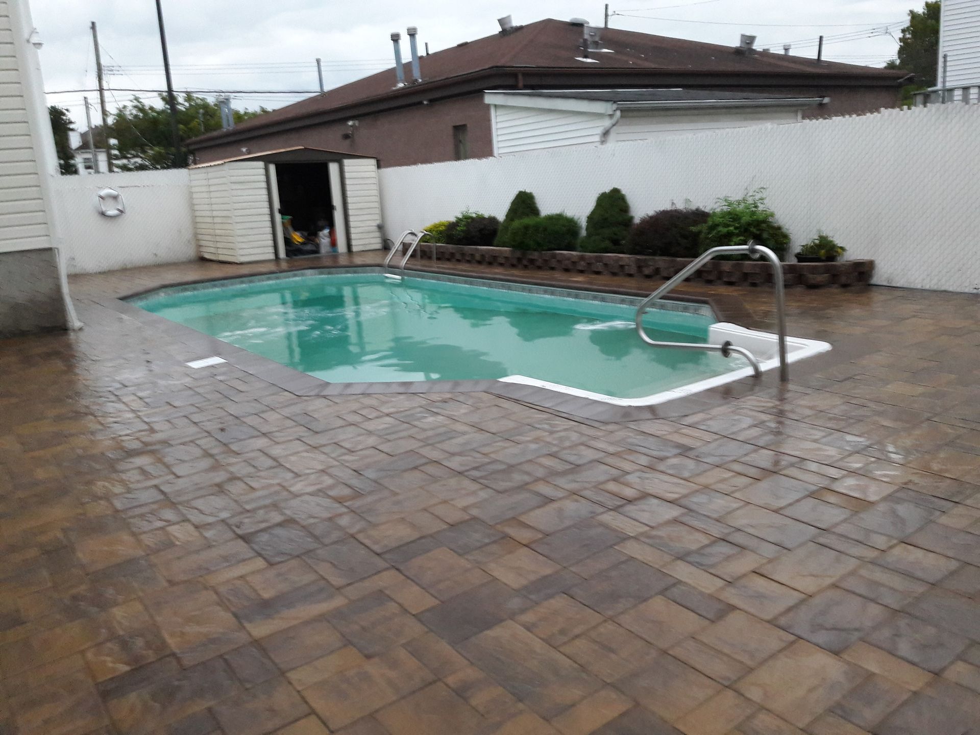 An outdoor rectangular swimming pool surrounded by a patterned brown stone paver patio, a white fence, and greenery.