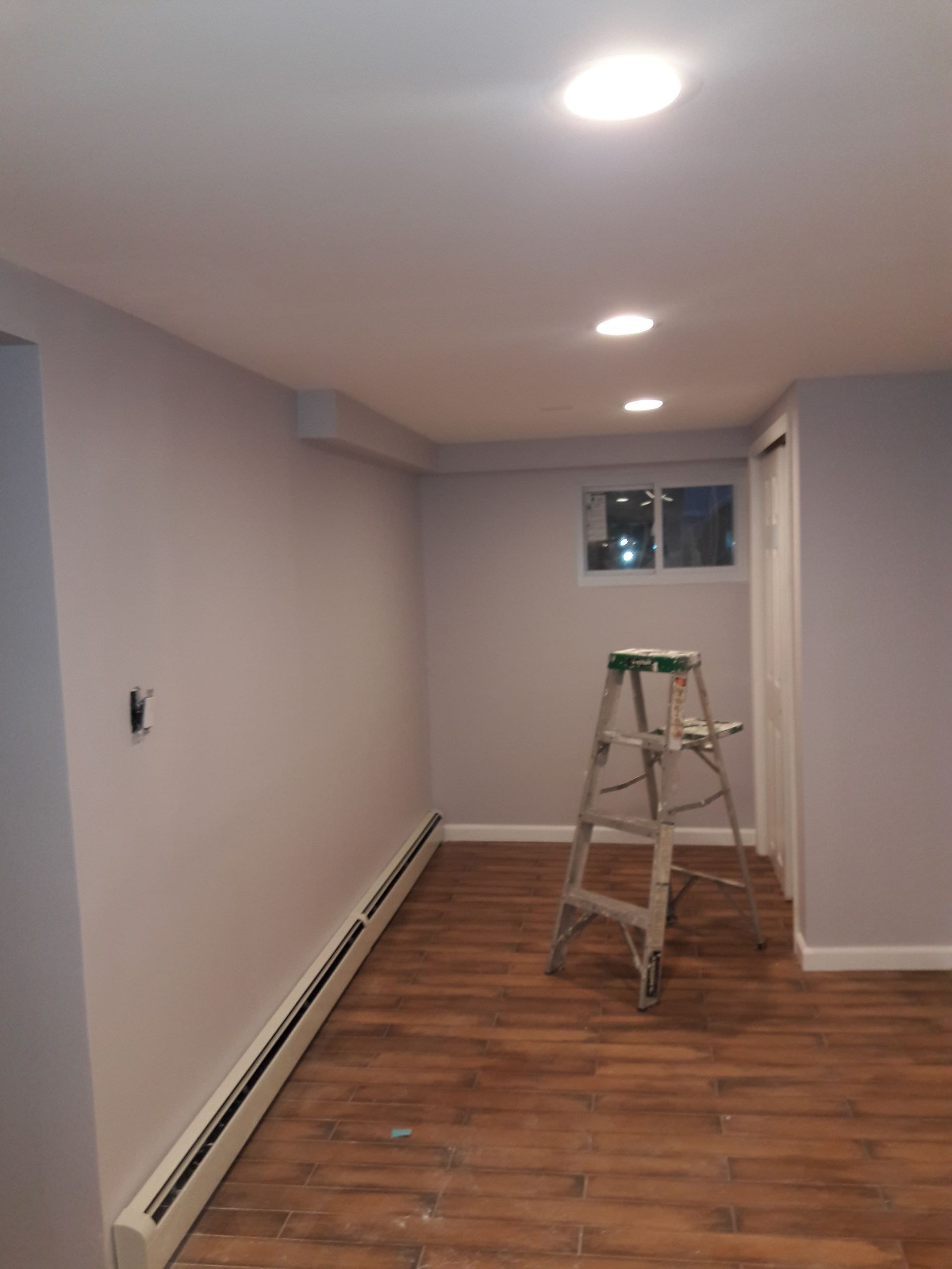 A finished basement room with grey walls, wooden flooring, recessed lighting, and a metal ladder standing in the center.