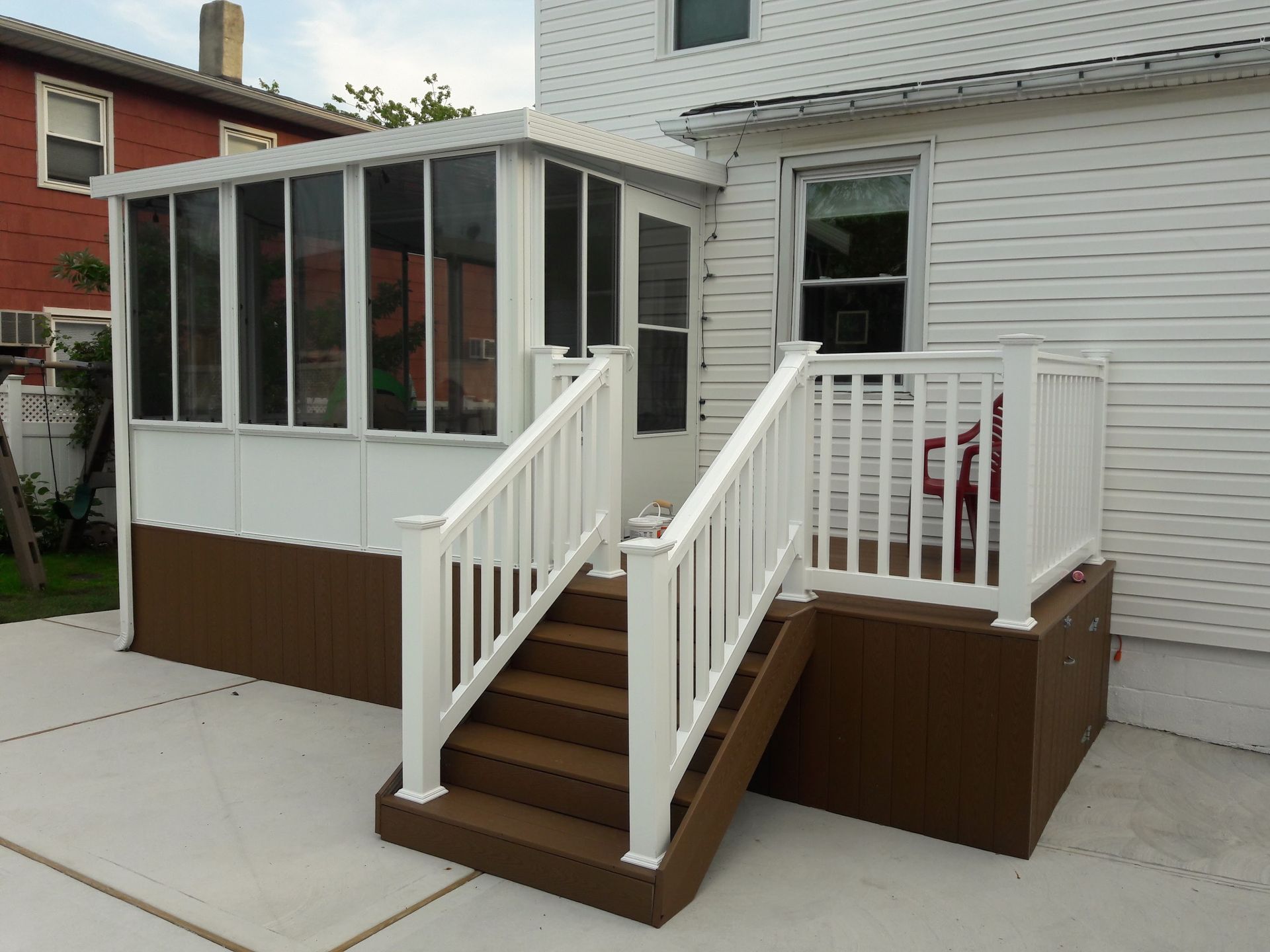 A white sunroom attached to a house, featuring brown skirting, a matching brown staircase, and white railings.