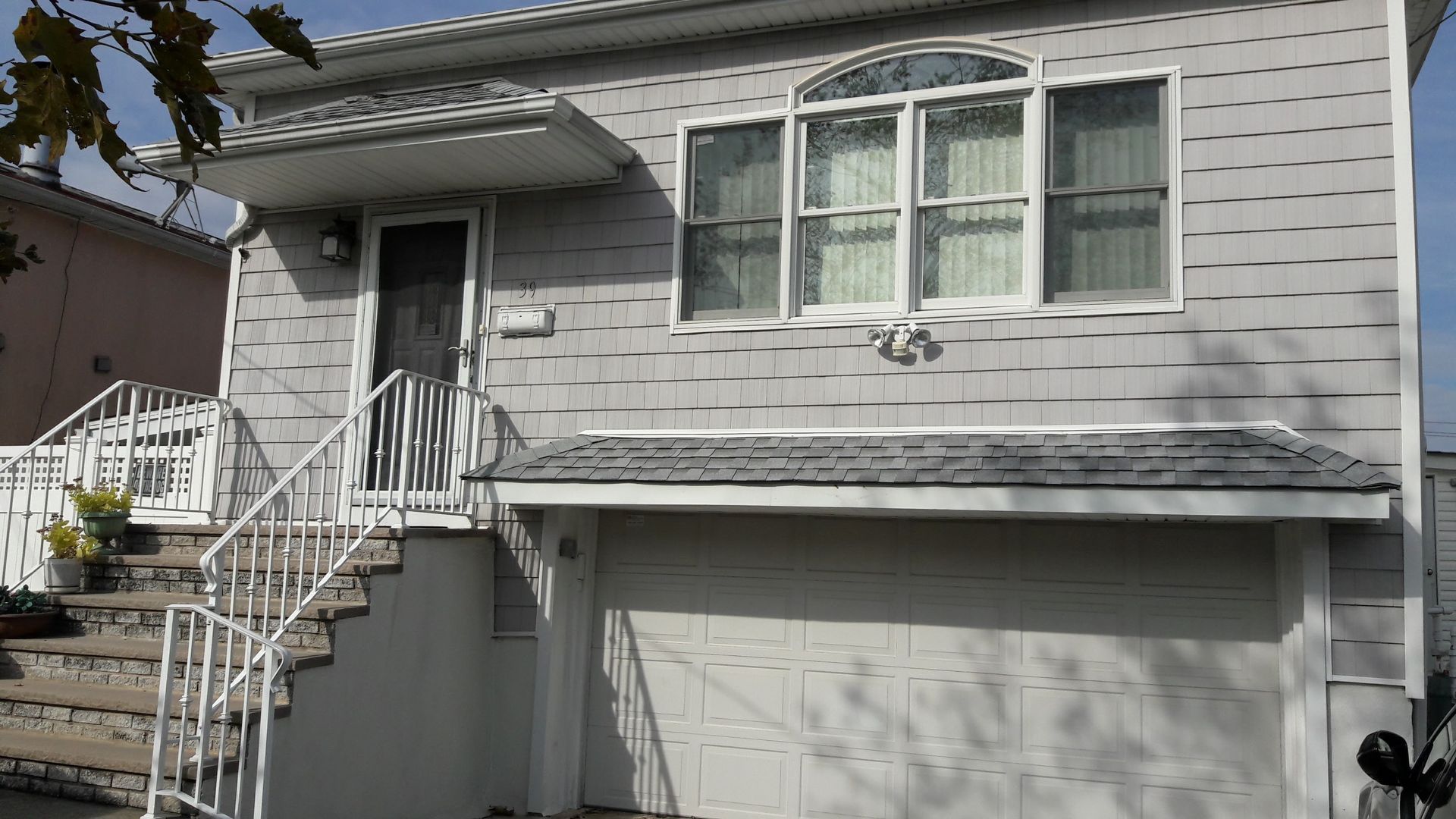 A two-story light gray house with a garage, stairs leading to the front entrance, and a large three-section window.