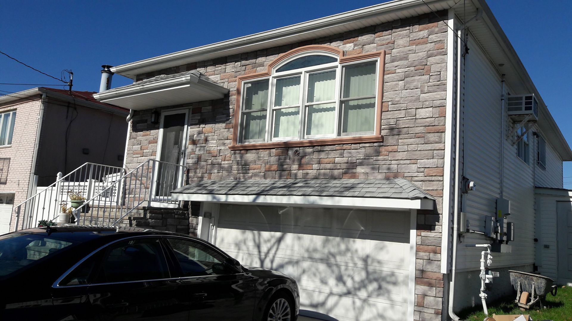 A two-story stone-faced house with a single-car garage and a front staircase leading to an entrance, under a blue sky.