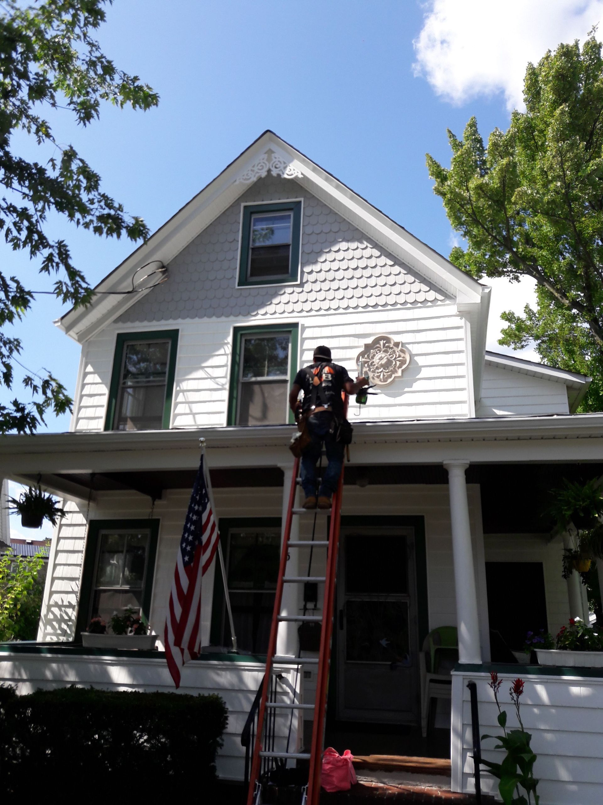 A person on a ladder working on the exterior of a white, two-story house with an American flag on the porch.