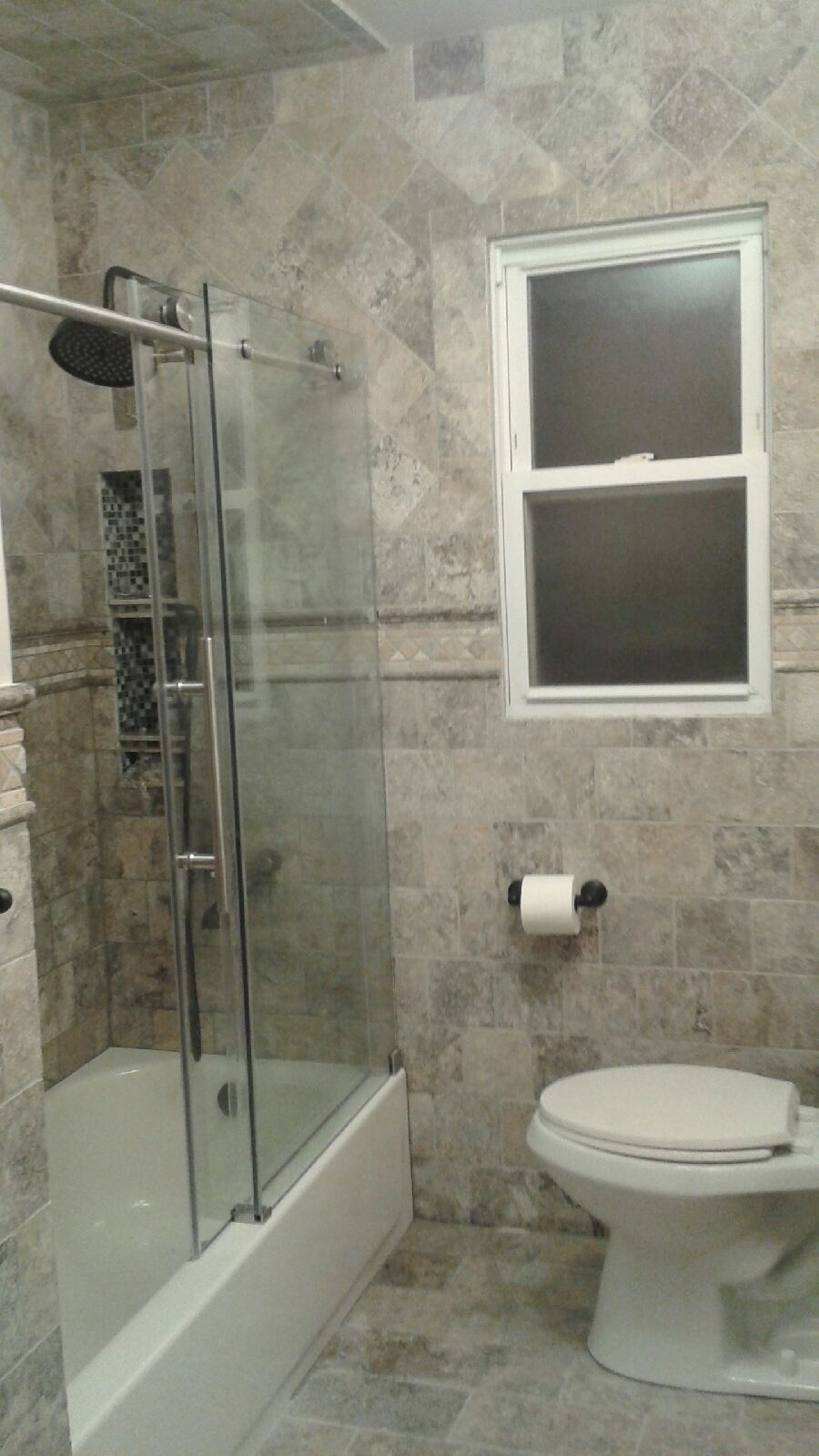 A bathroom featuring a glass-enclosed shower, a white toilet, and tan stone-tiled walls and floors.