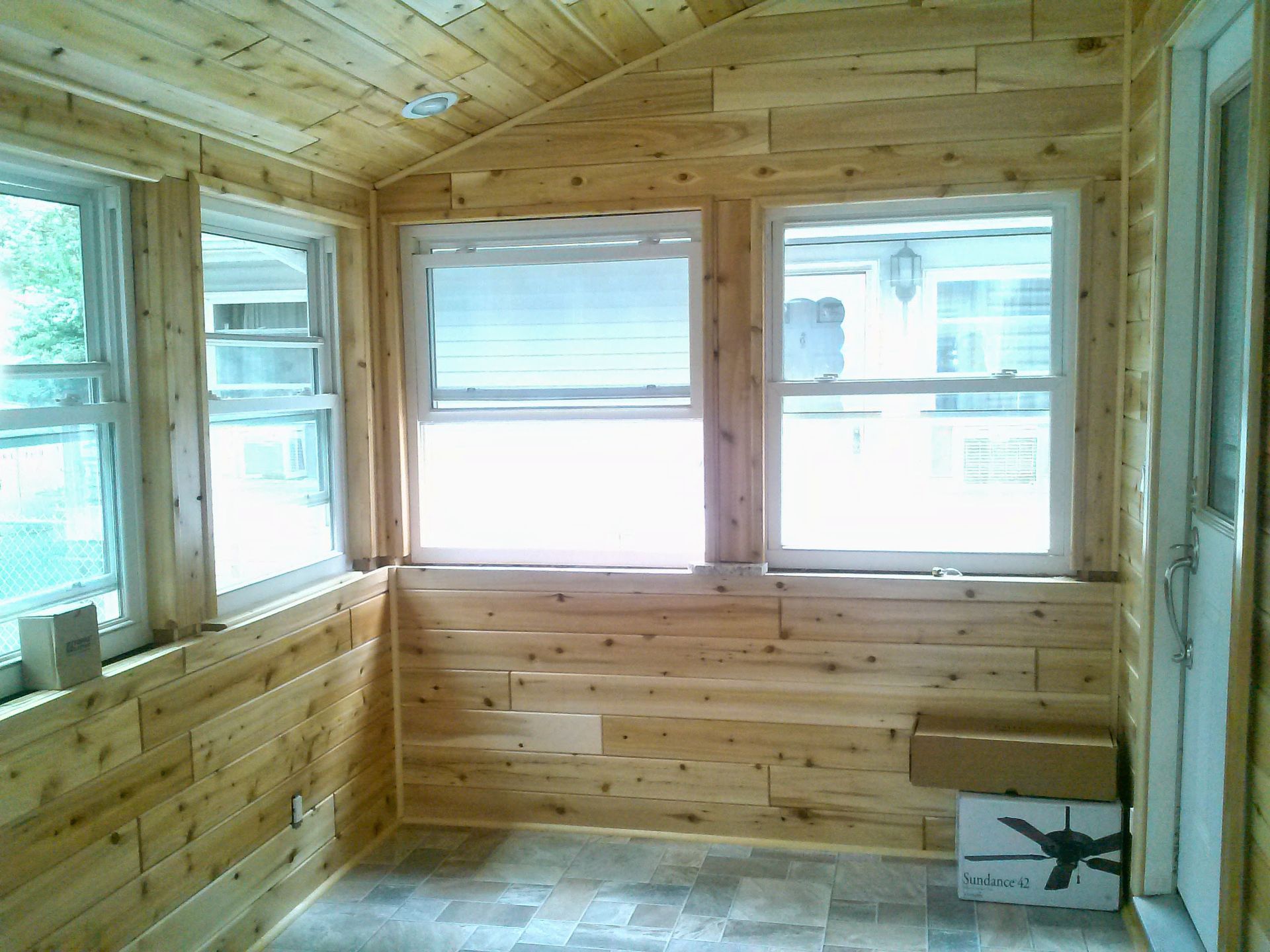 A bright, unfinished sunroom featuring light-colored wood paneled walls, a vaulted ceiling, multiple windows, and flooring.
