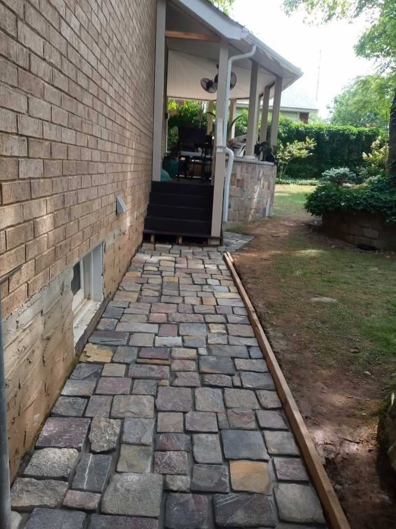 A stone paver walkway runs along a brick house wall, leading to a wooden deck with steps on a sunny day.