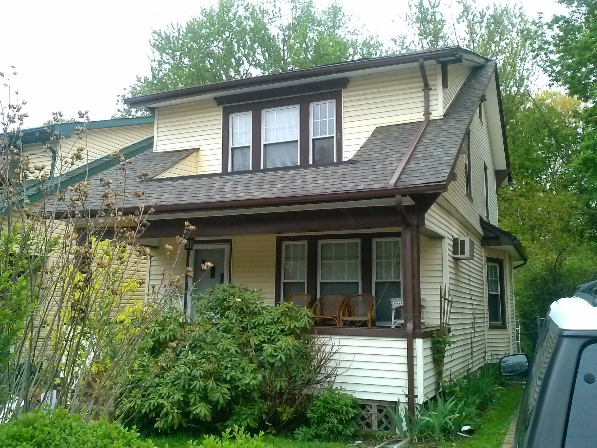 A two-story light yellow house with dark brown trim, a front porch, and a dormer window, surrounded by trees and greenery.