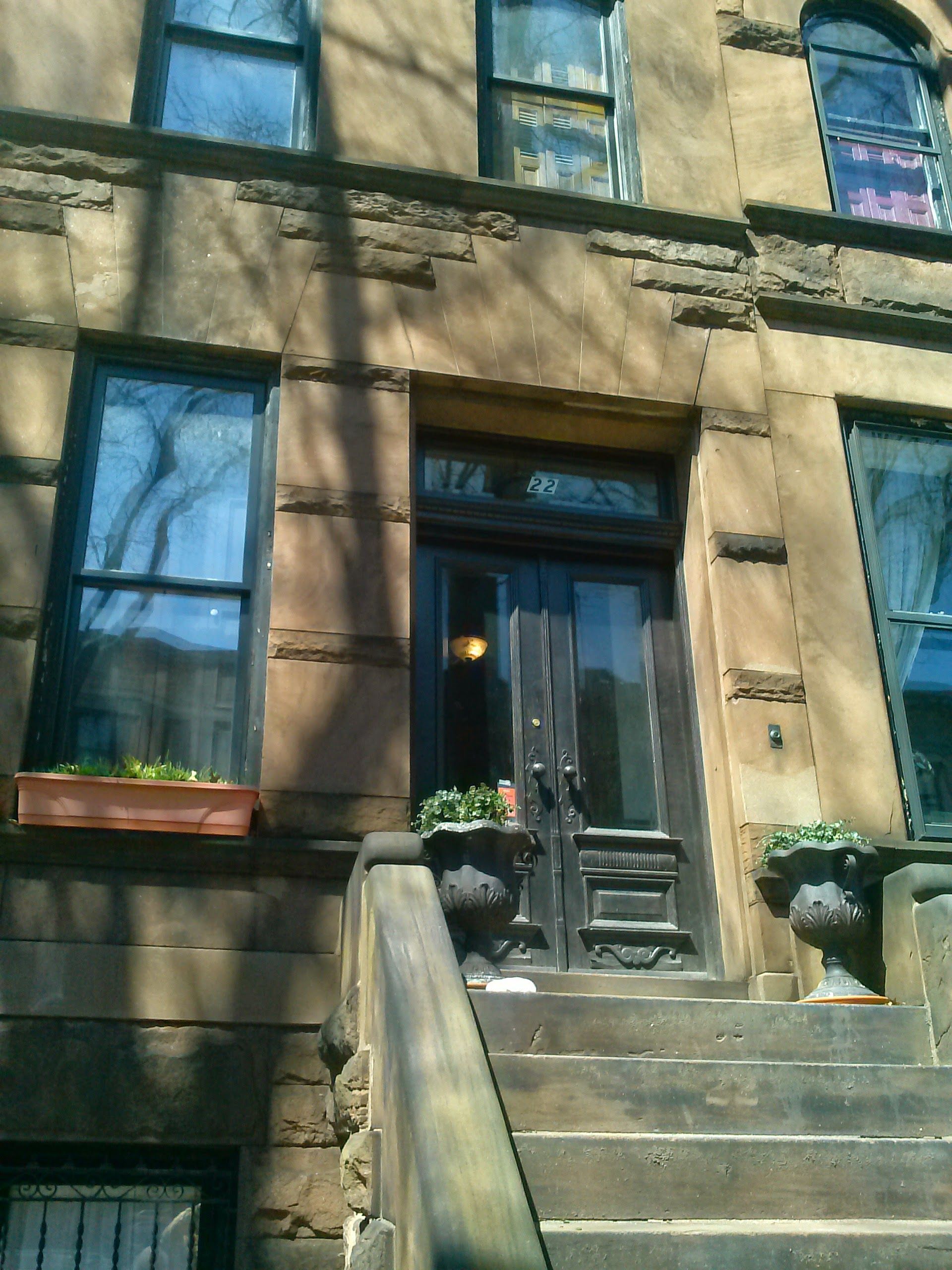 The stone entrance of a brownstone building with steps, a black double door, and windows with planters.