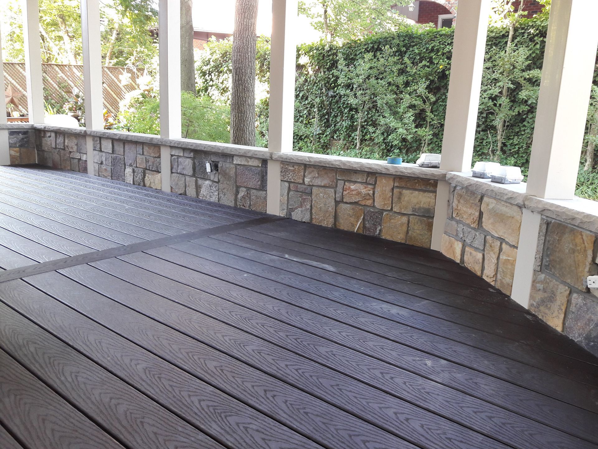 A dark wooden deck with a low stone retaining wall and white support pillars, overlooking a lush green yard.