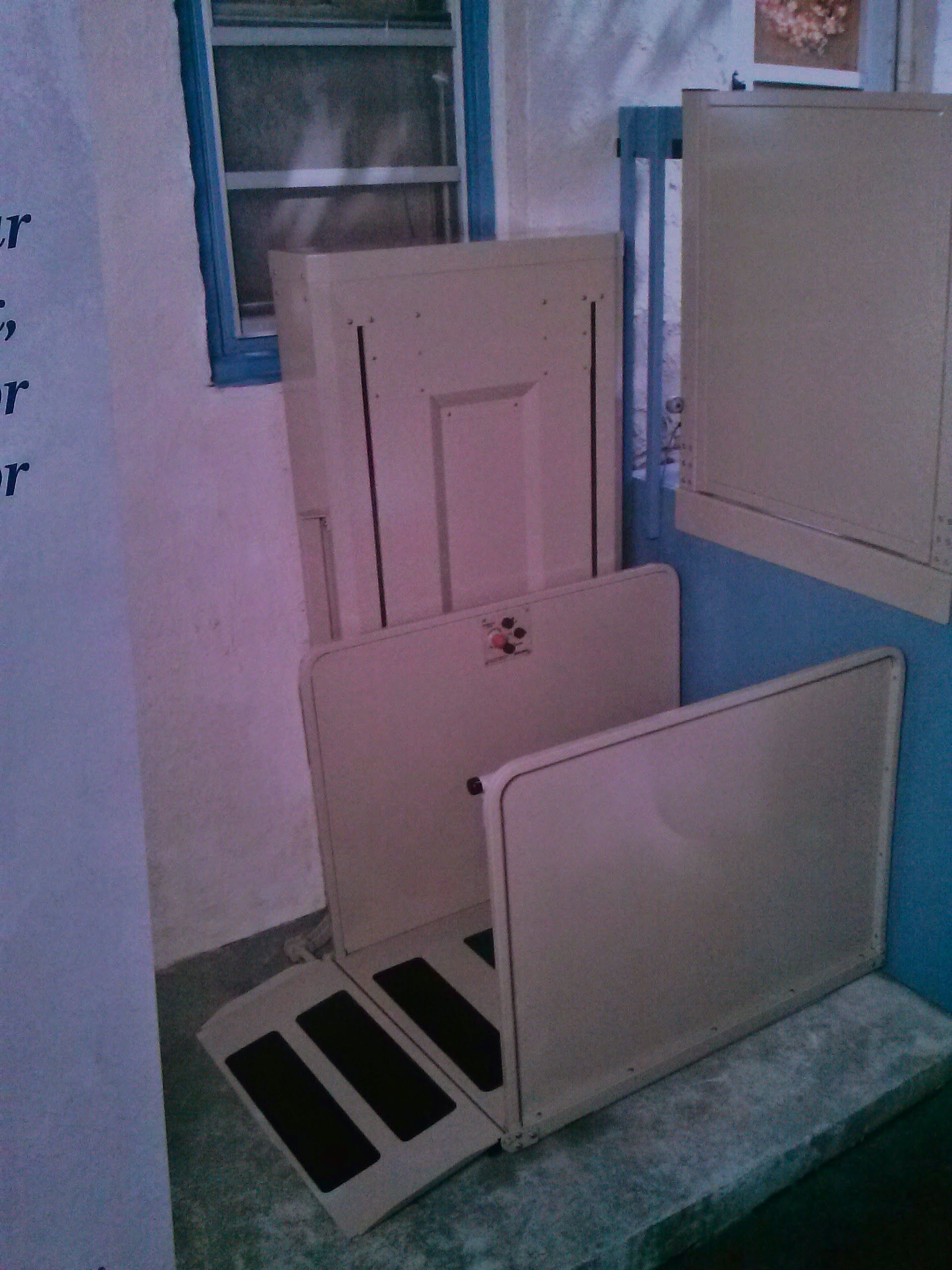 A light-tan wheelchair platform lift installed on a concrete porch near a window and a door.