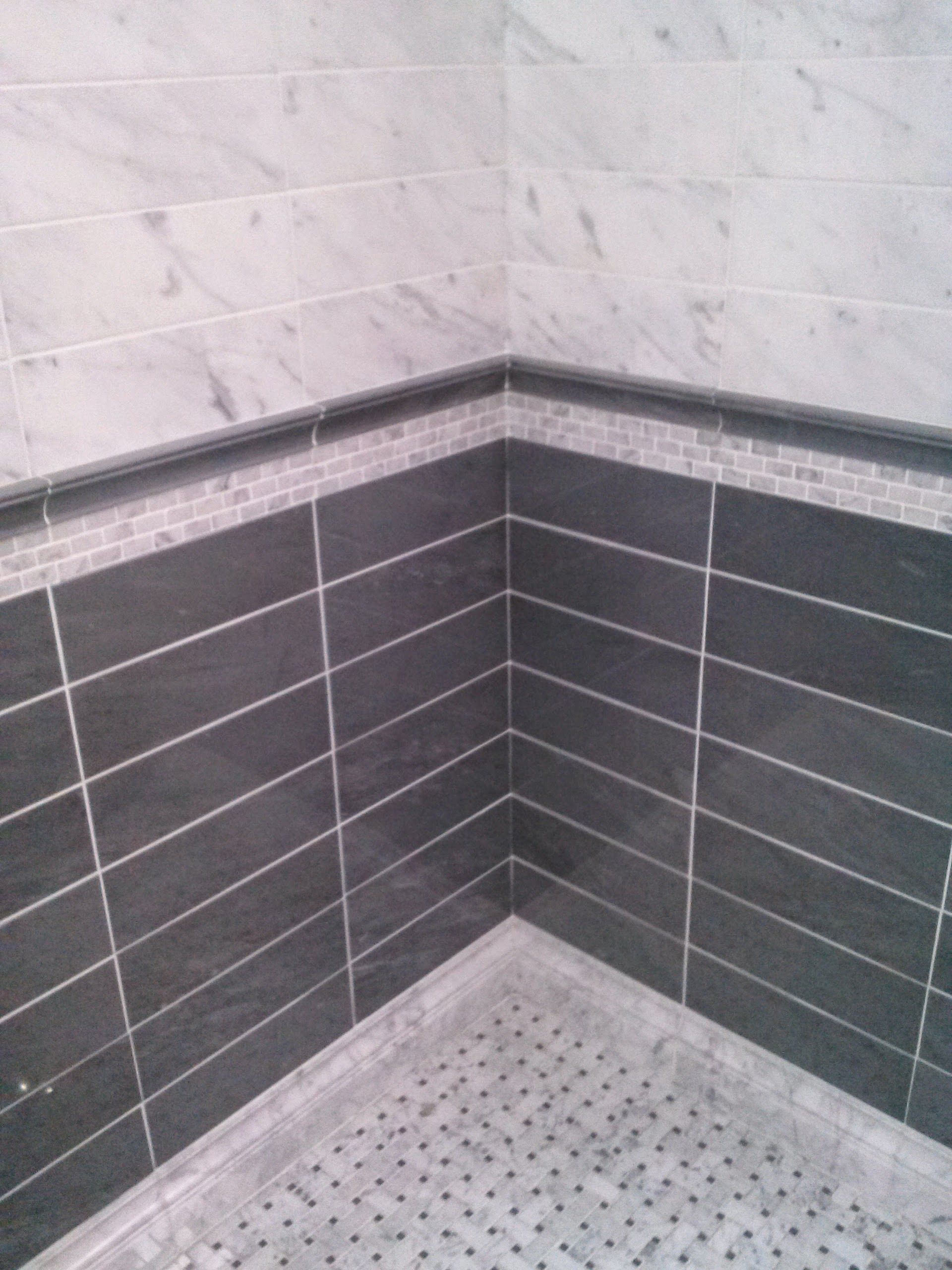 A corner of a shower stall with white marble upper tiles, a dark horizontal accent border, and dark grey lower tiles.