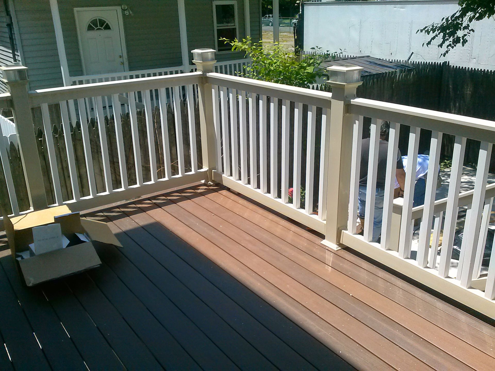 A wooden deck with white railings featuring two different-colored boards, partially shaded, with a cardboard box nearby.