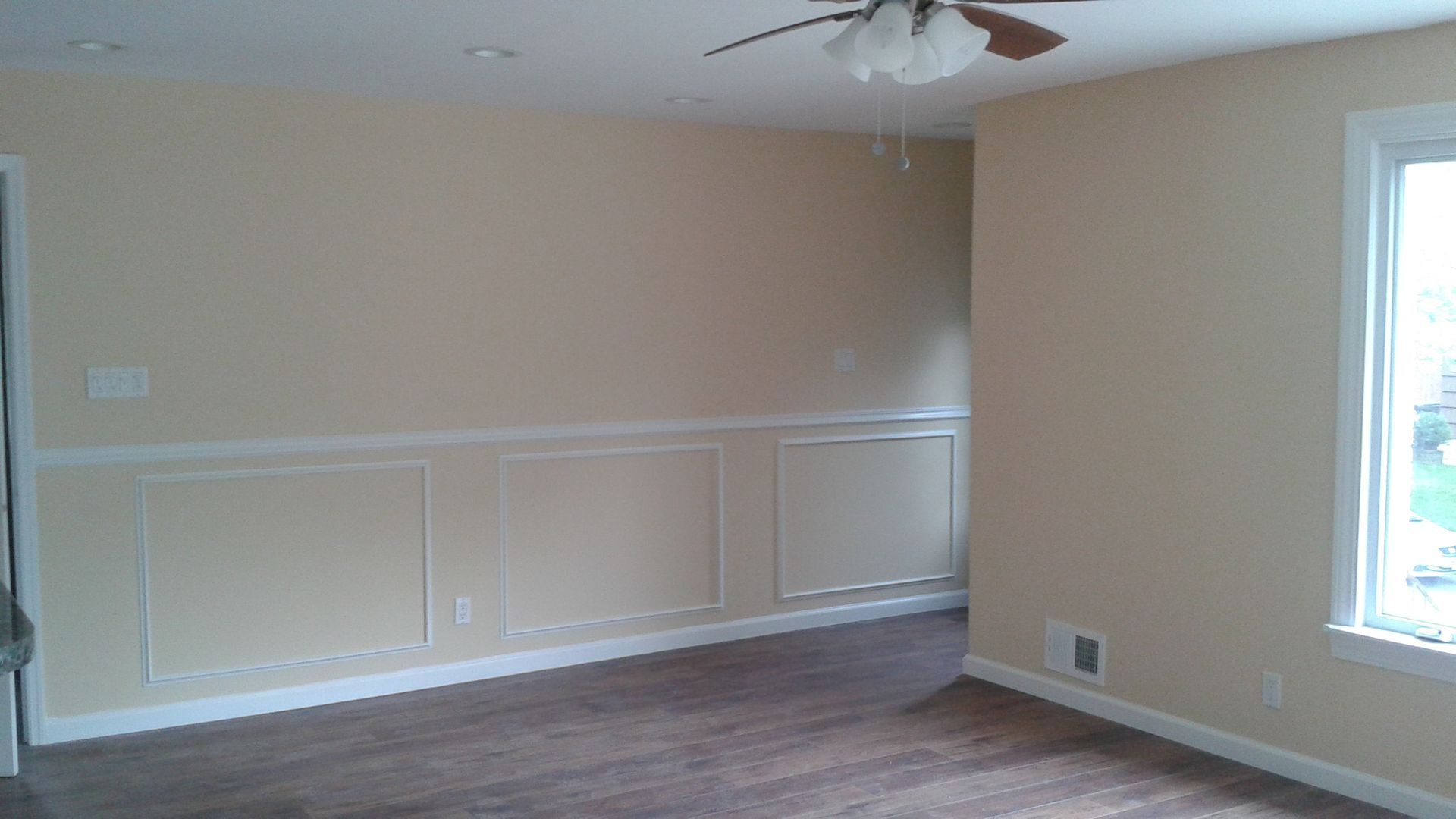 A beige room with white wainscoting, wood-look flooring, a ceiling fan, and a window on the right wall.