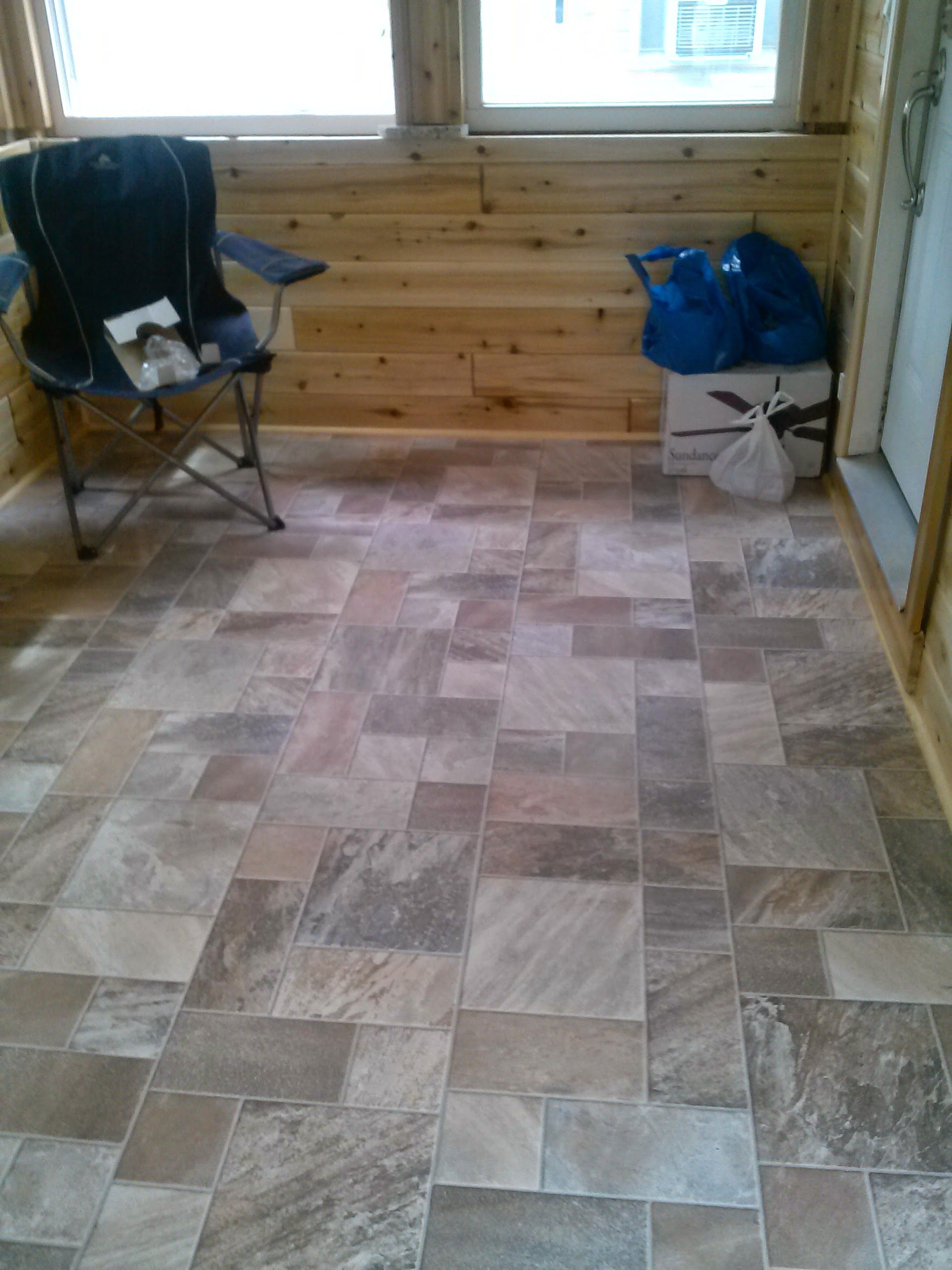 A room with natural wood walls and a floor tiled in a multi-sized, earth-toned stone pattern, with a chair and bags inside.