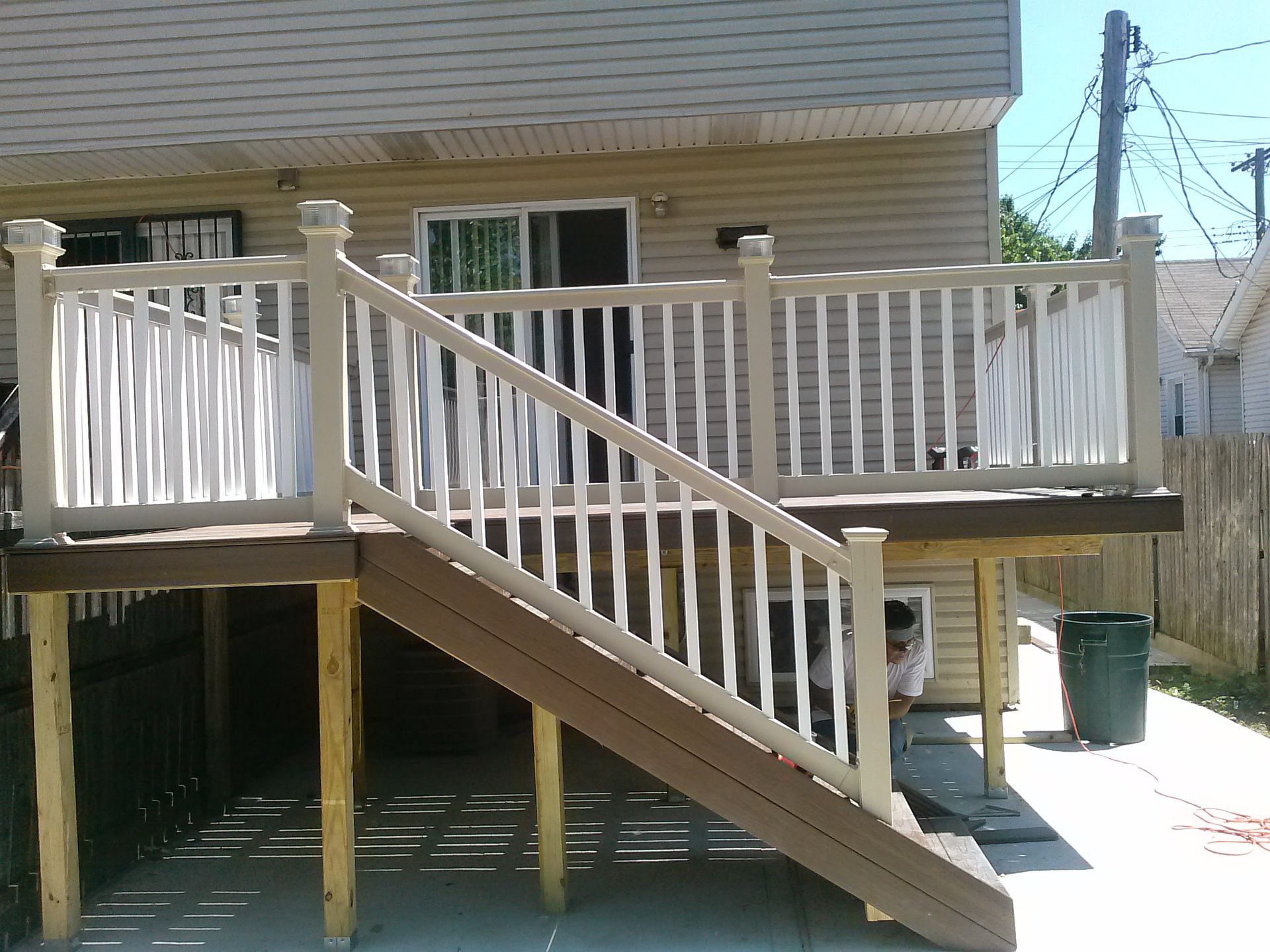 A newly built brown wooden deck with white railings and stairs, positioned at the back of a house over a concrete patio.