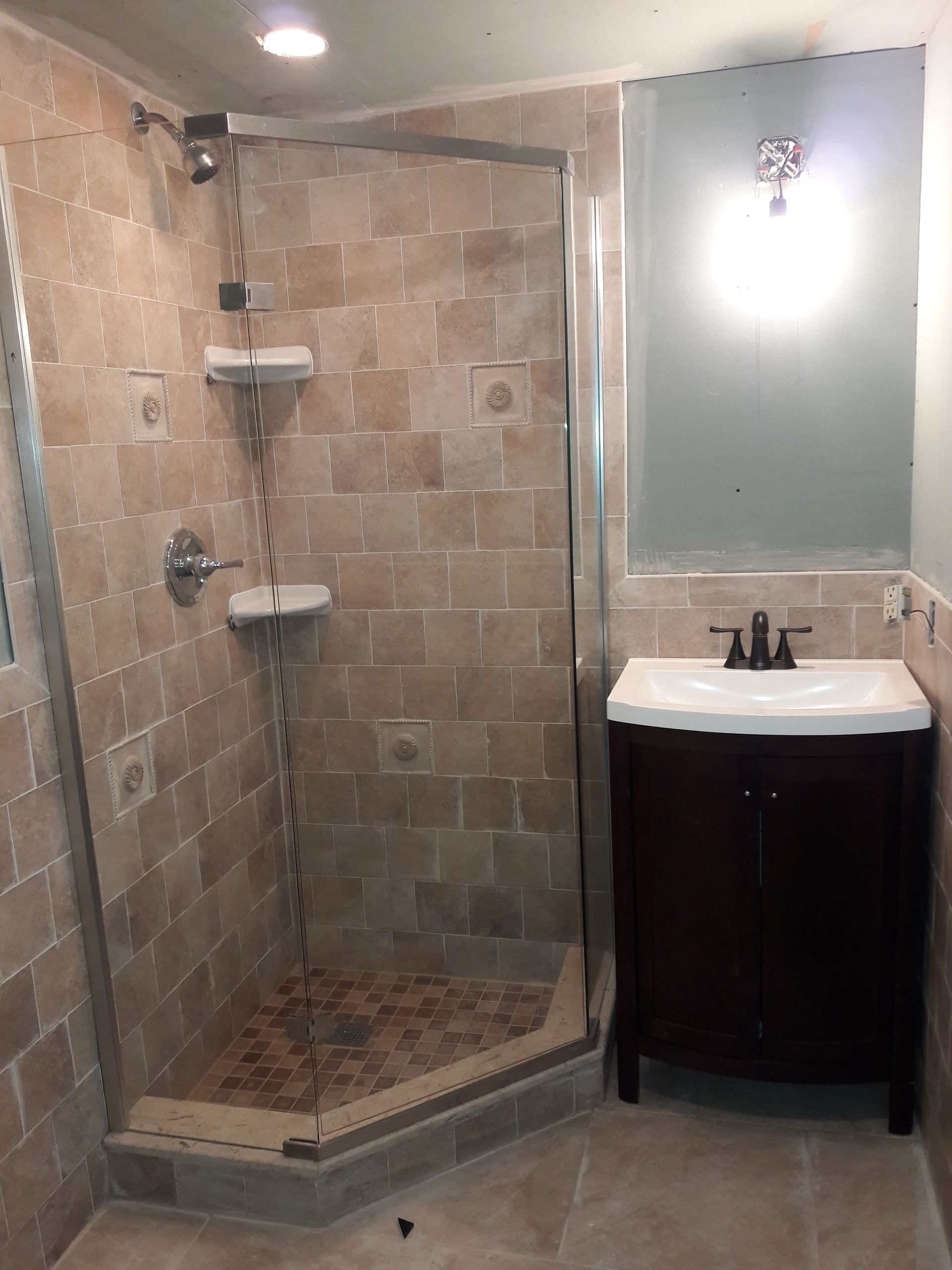 A tiled corner shower stall with a glass door next to a small dark wood vanity with a white sink and a large mirror.