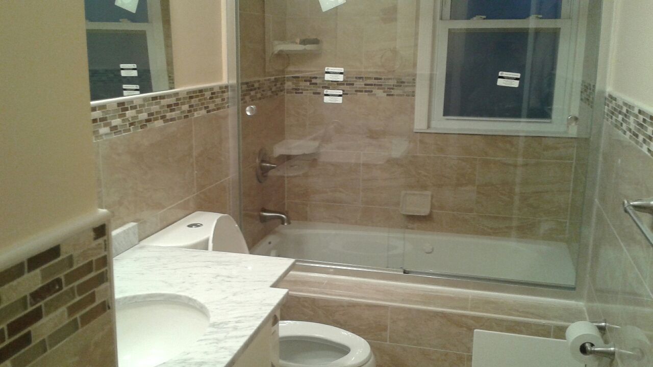 A renovated bathroom featuring a marble-topped vanity, a toilet, and a tiled bathtub with a glass door and window.