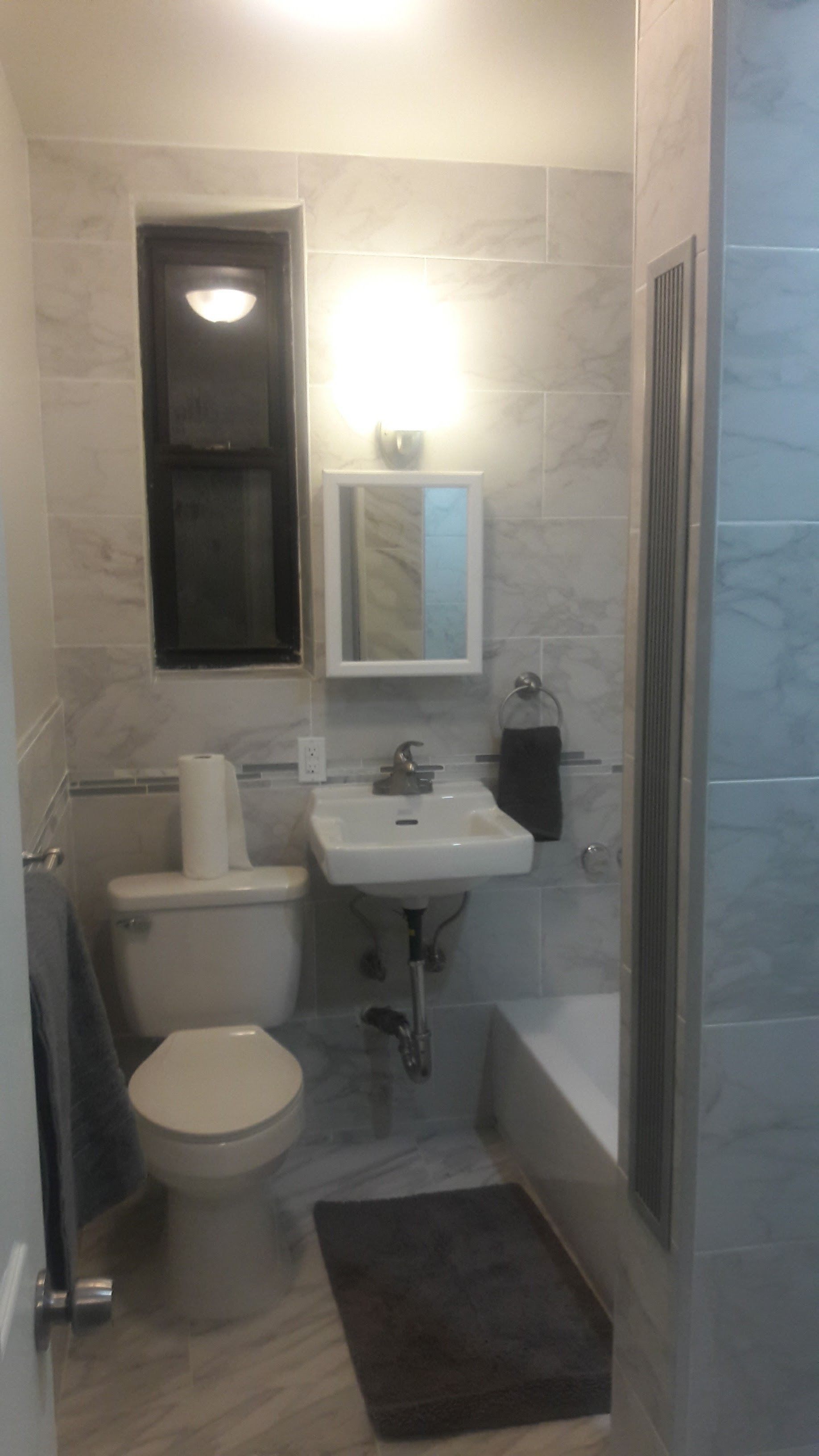 A small, bright bathroom with light grey marble-style tiled walls, a white toilet, a wall-mounted sink, and a shower area.
