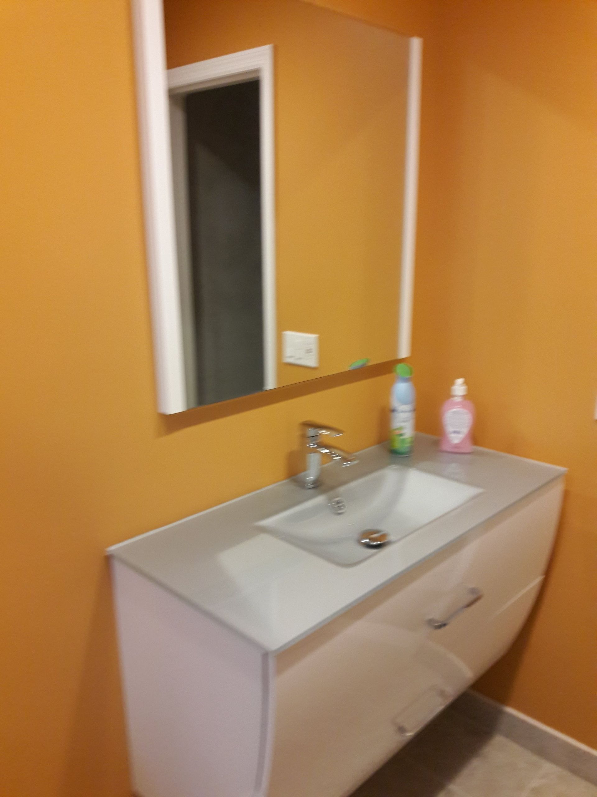 A modern white bathroom vanity with a silver faucet and mirror against orange walls, featuring soap bottles on the counter.