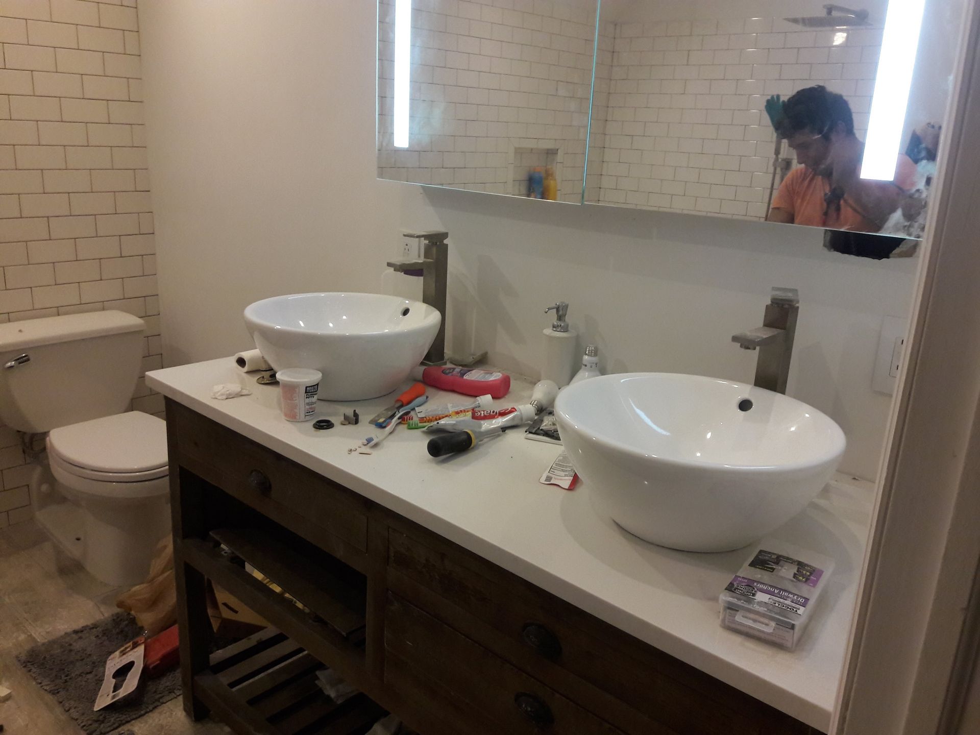 A double vanity bathroom with white vessel sinks, a wood cabinet, and a toilet, with tools scattered on the countertop.