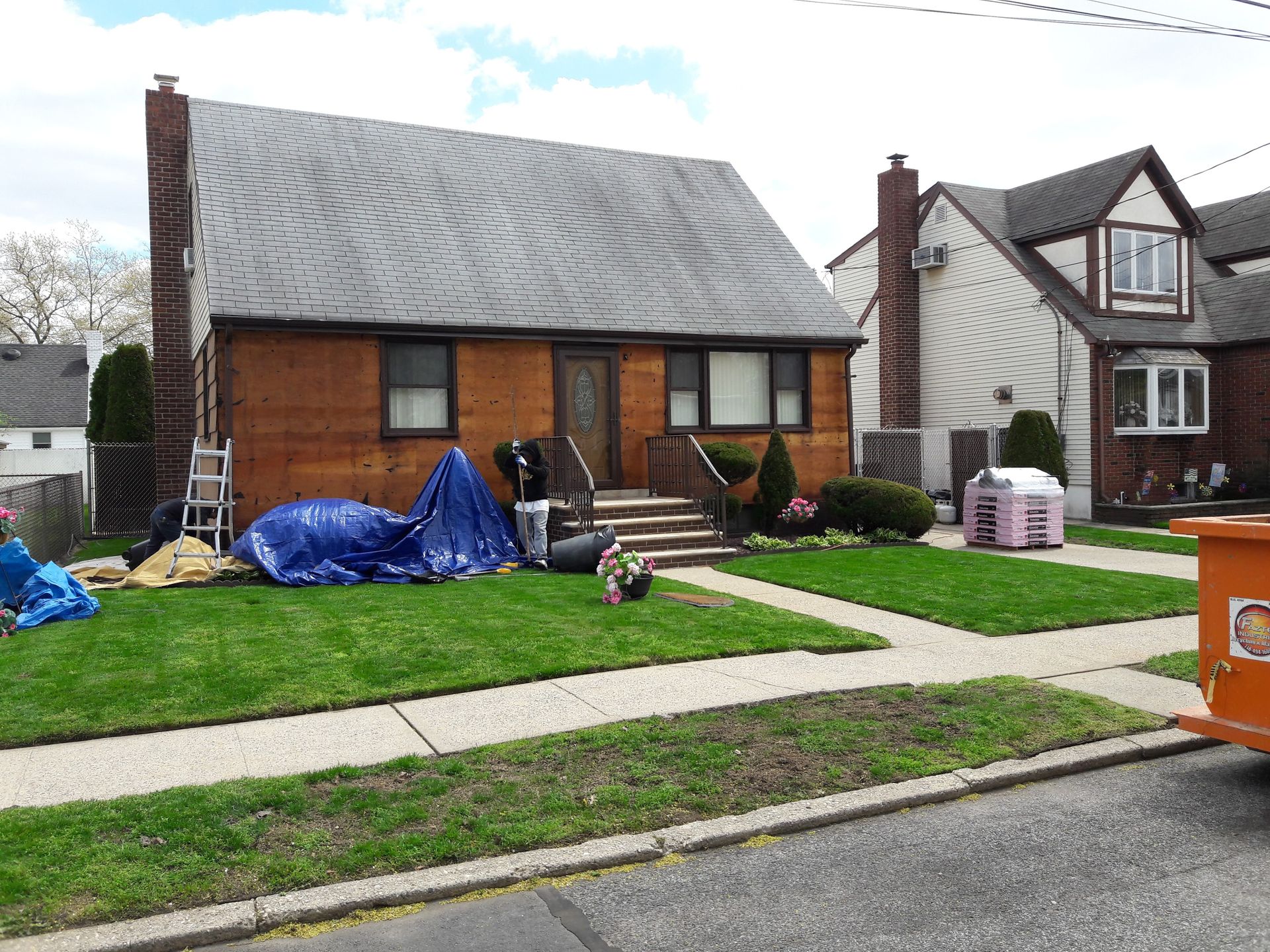 A residential house with wood siding and a steep roof, featuring a blue tarp on the lawn and a dumpster nearby.