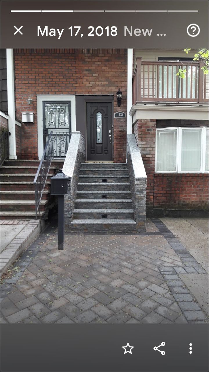 A residential brick building entrance with a set of stone steps leading to a dark door and a gray brick paved walkway.