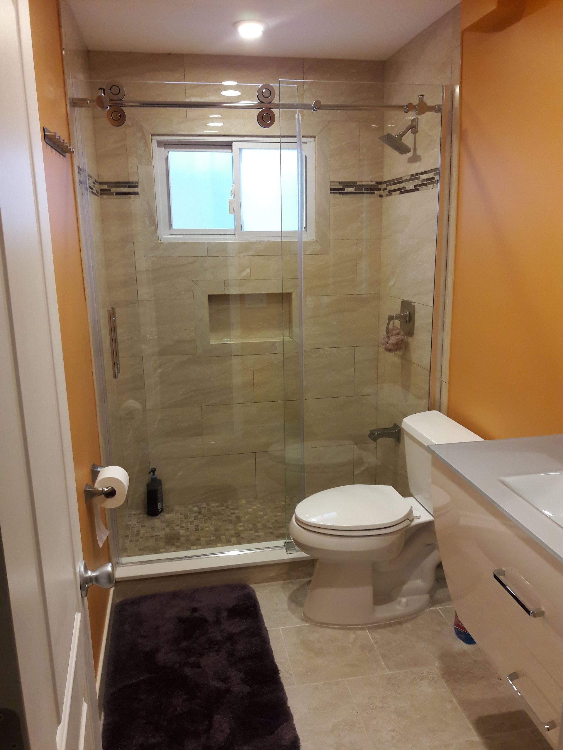 A bathroom featuring an orange wall, a white vanity, a toilet, and a glass-enclosed shower with tiled walls and flooring.