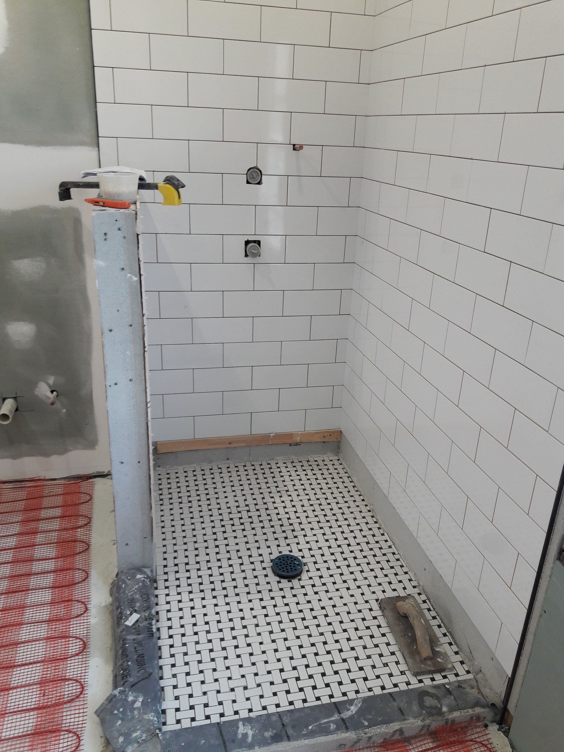 A shower stall under construction with white brick-style wall tiles, a mosaic-tiled floor, and exposed plumbing fixtures.
