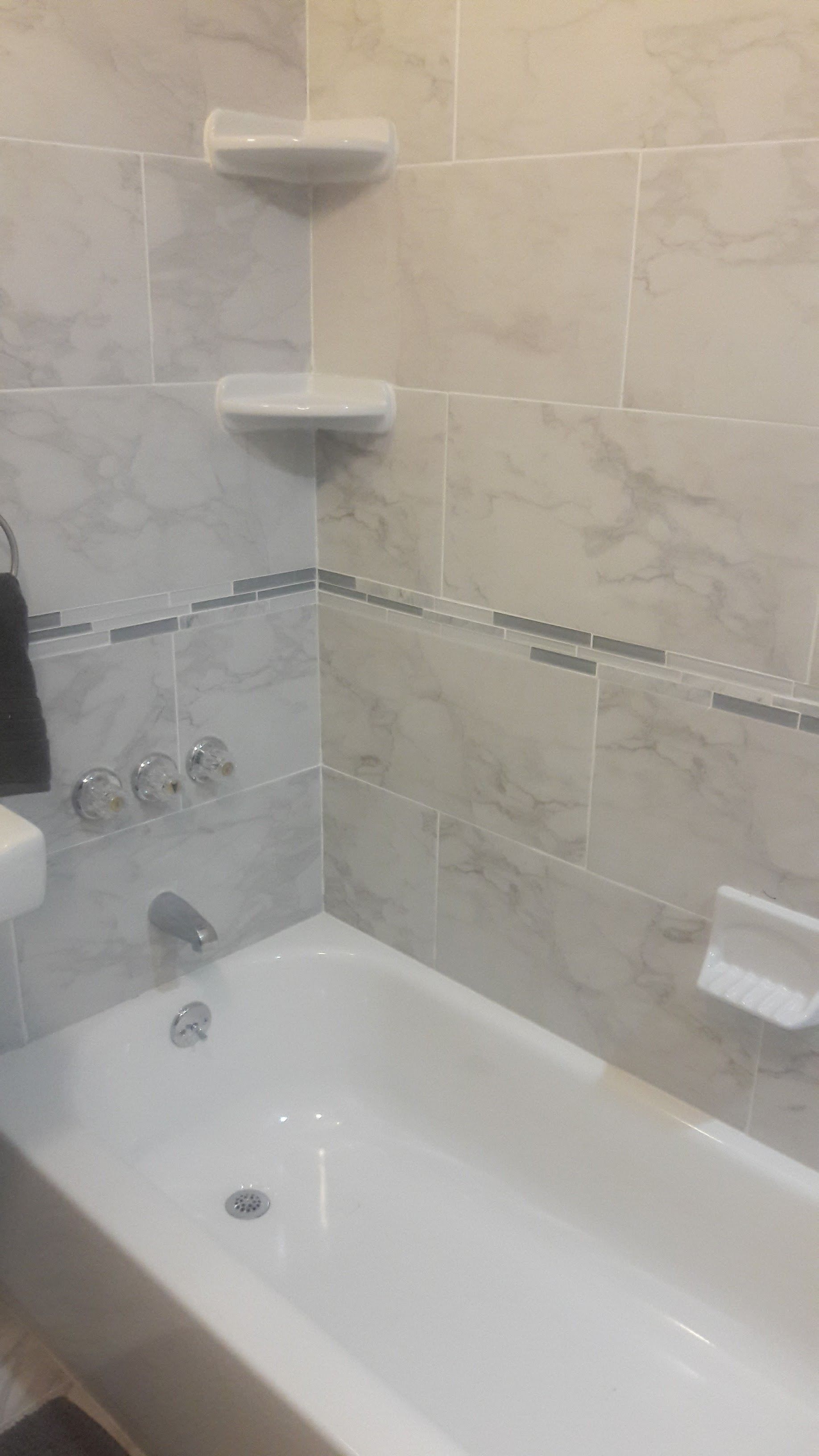 A white bathtub and shower surround with beige marble-patterned tiles, two built-in corner shelves, and chrome fixtures.