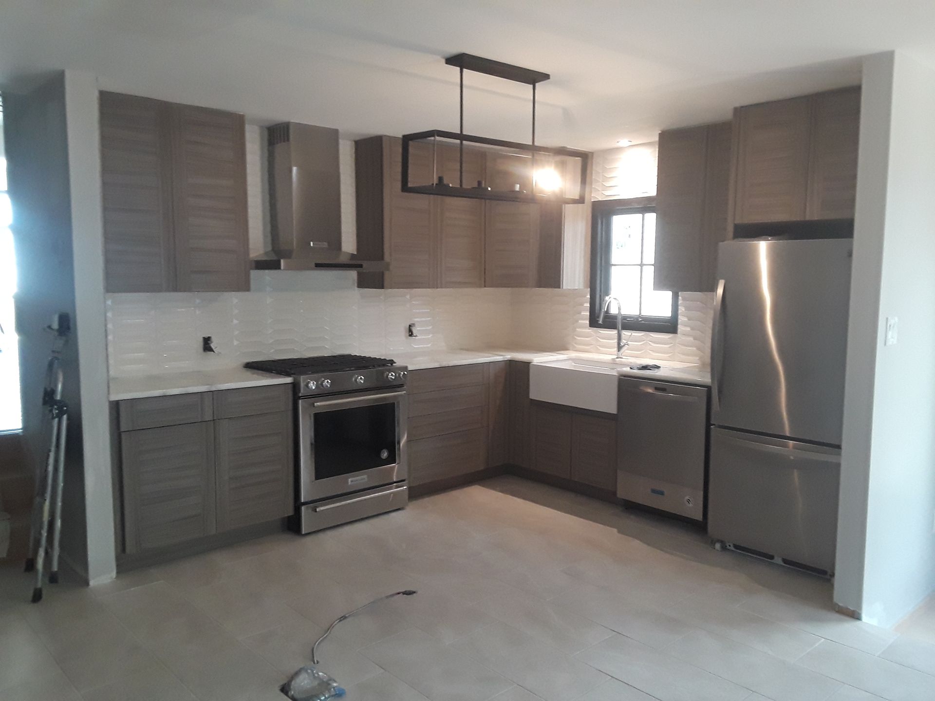 A modern kitchen with grey cabinets, stainless steel appliances, a white farmhouse sink, and a rectangular pendant light.