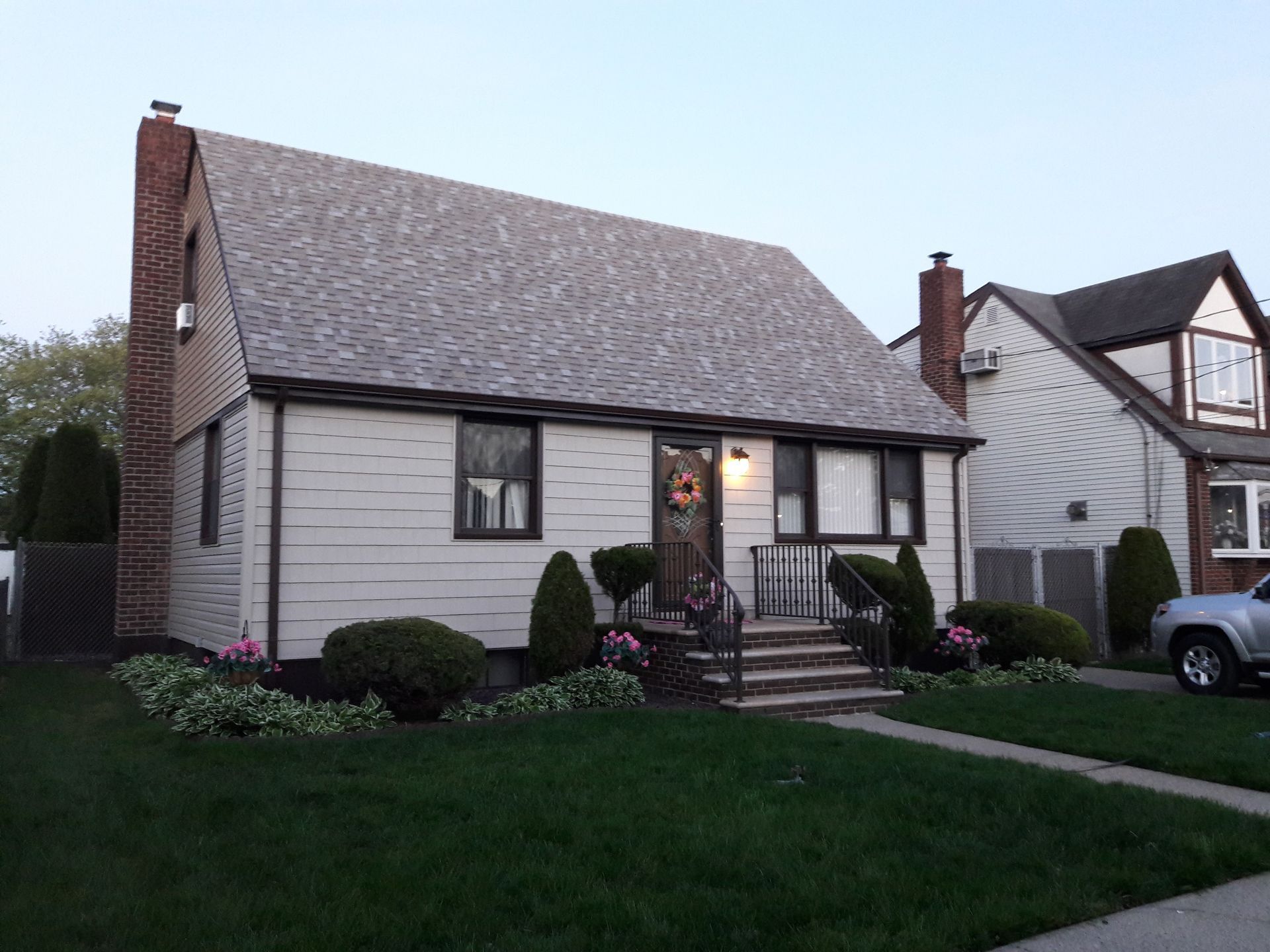 A single-story, light-colored house with a peaked roof, brick chimney, and a small front yard with shrubs.