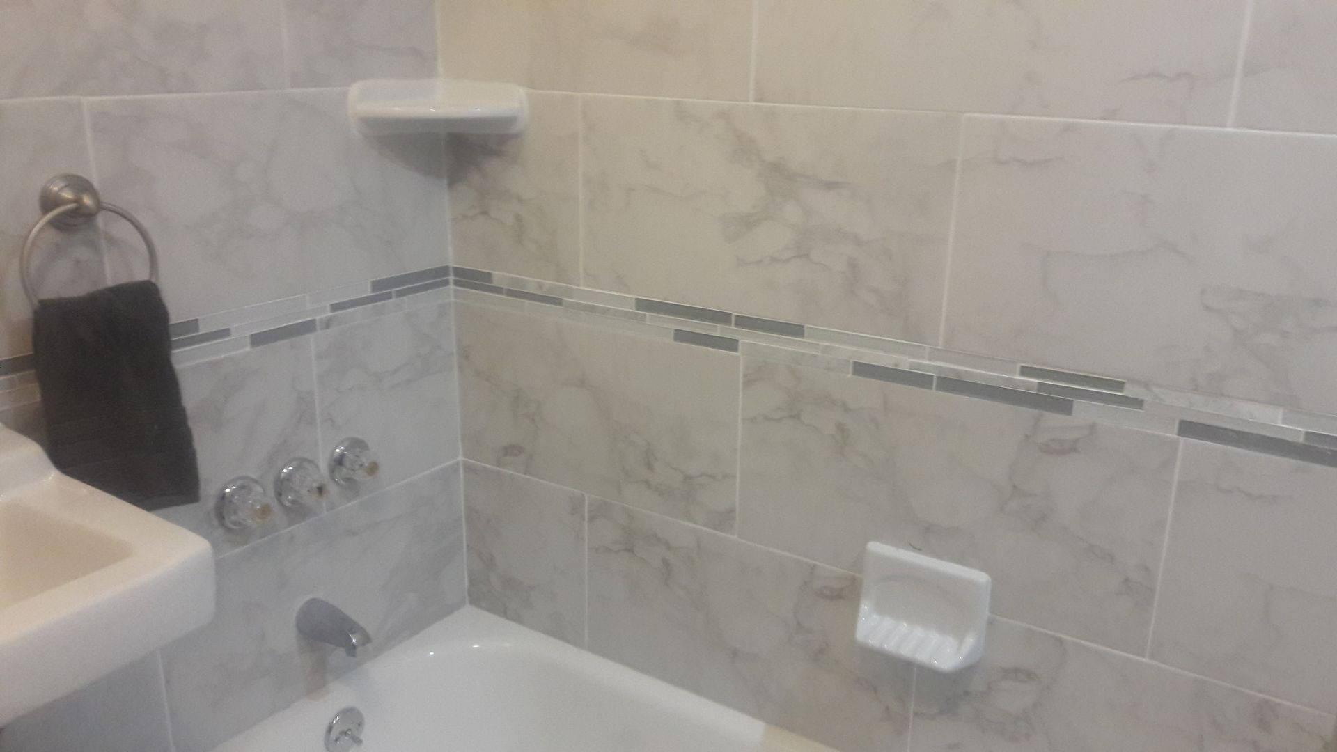 A bathroom tub and wall area with light gray marble tiles, a dark towel on a ring, and built-in soap dishes.