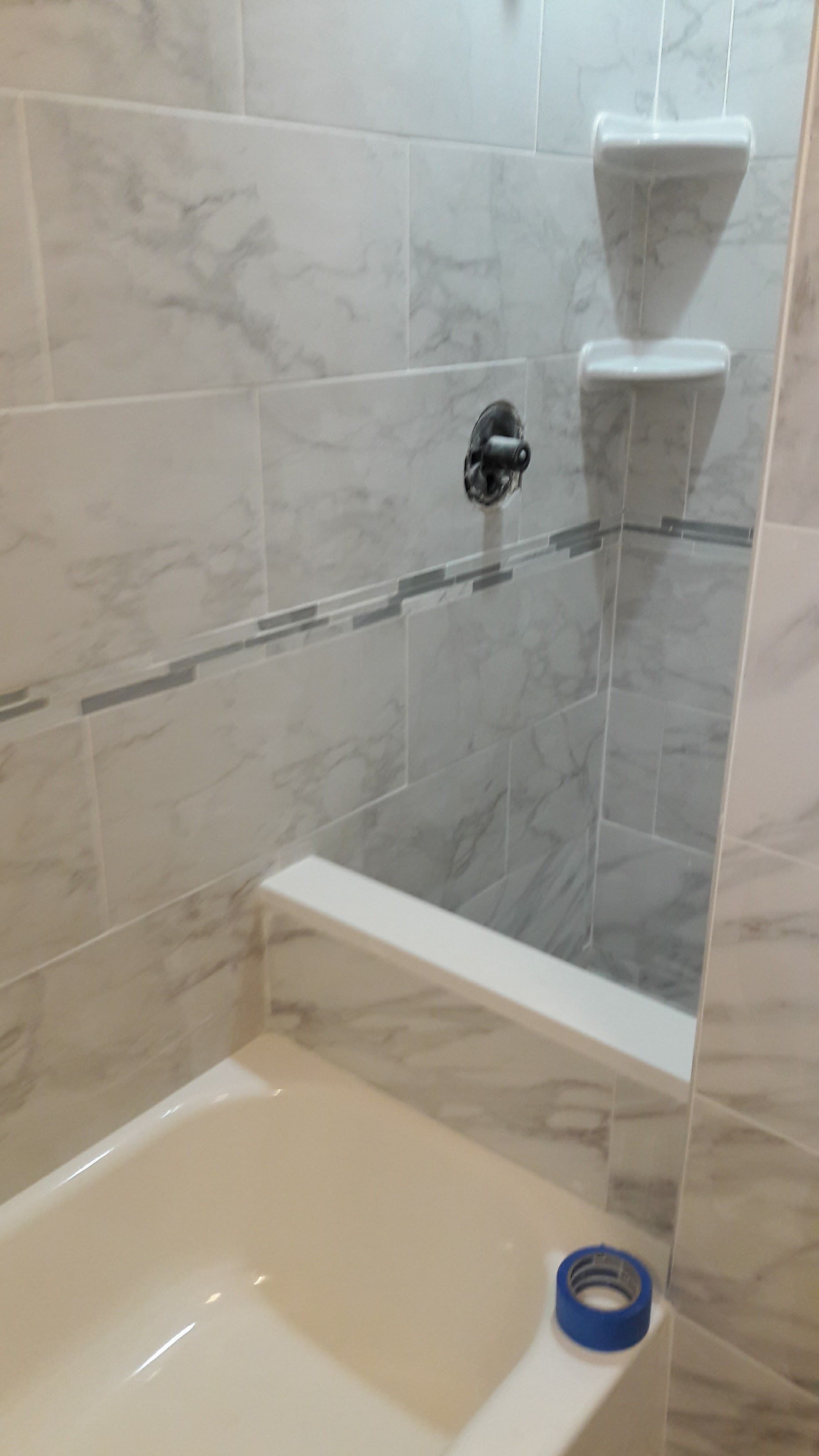 A tiled shower alcove featuring light grey marble-patterned walls, two built-in corner shelves, a shower faucet, and tape.