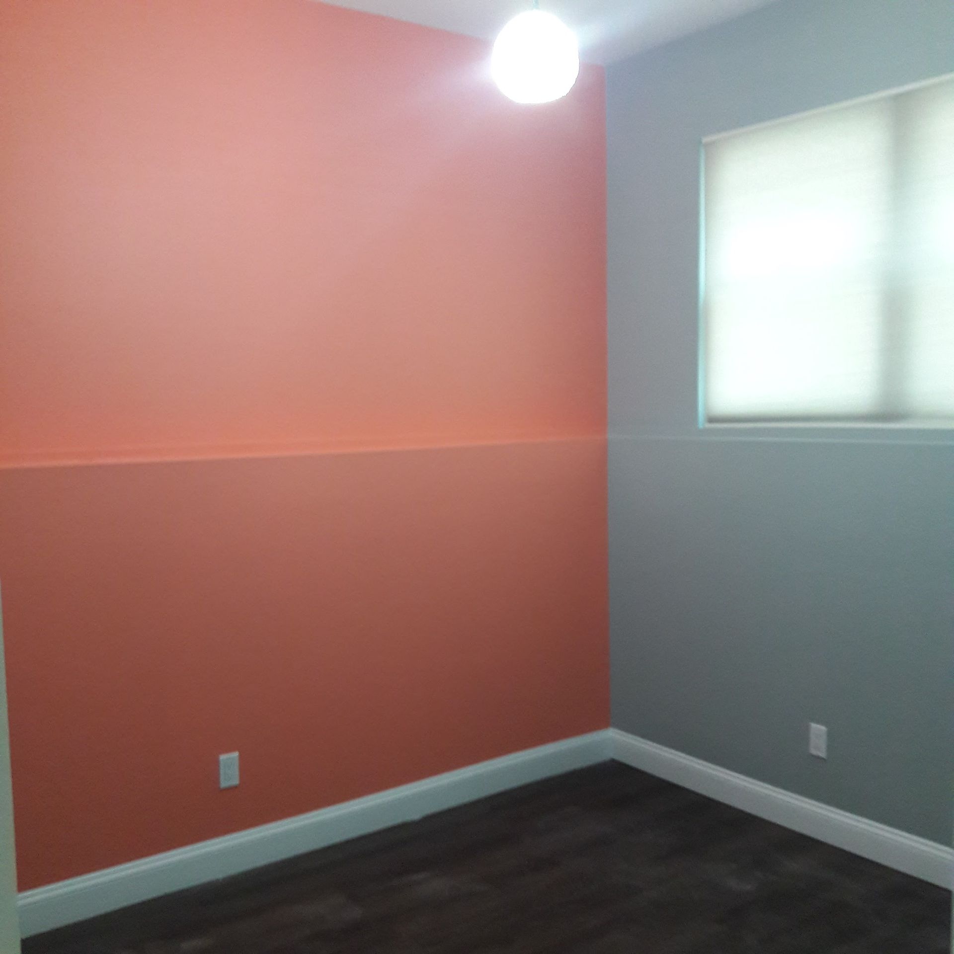 A room corner with two-toned peach walls and a gray wall featuring a window with a white shade, plus dark flooring.