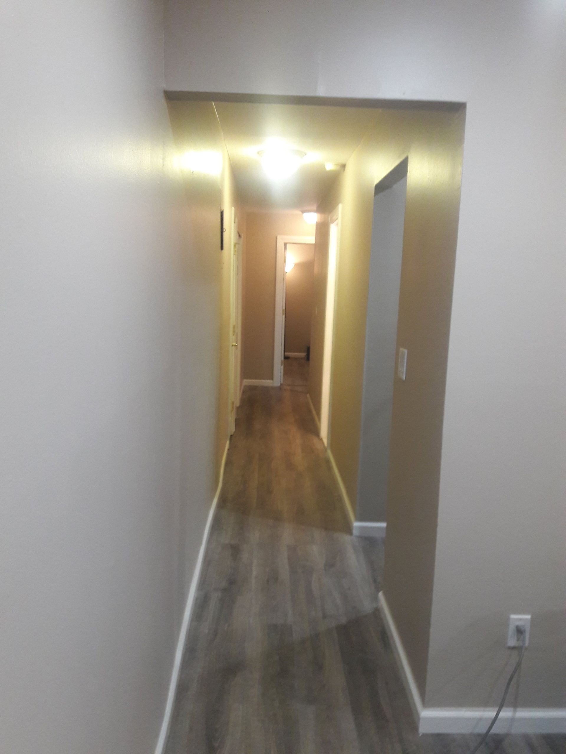 A narrow, well-lit indoor hallway with light brown walls and light gray wood-look flooring, leading to a distant doorway.