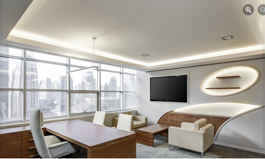 A modern office with a wooden desk and chair, a television on the wall, and seating area in front of large windows.