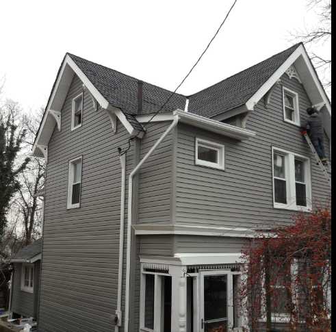 A person on a ladder repairs the exterior of a gray, two-story house with white trim.