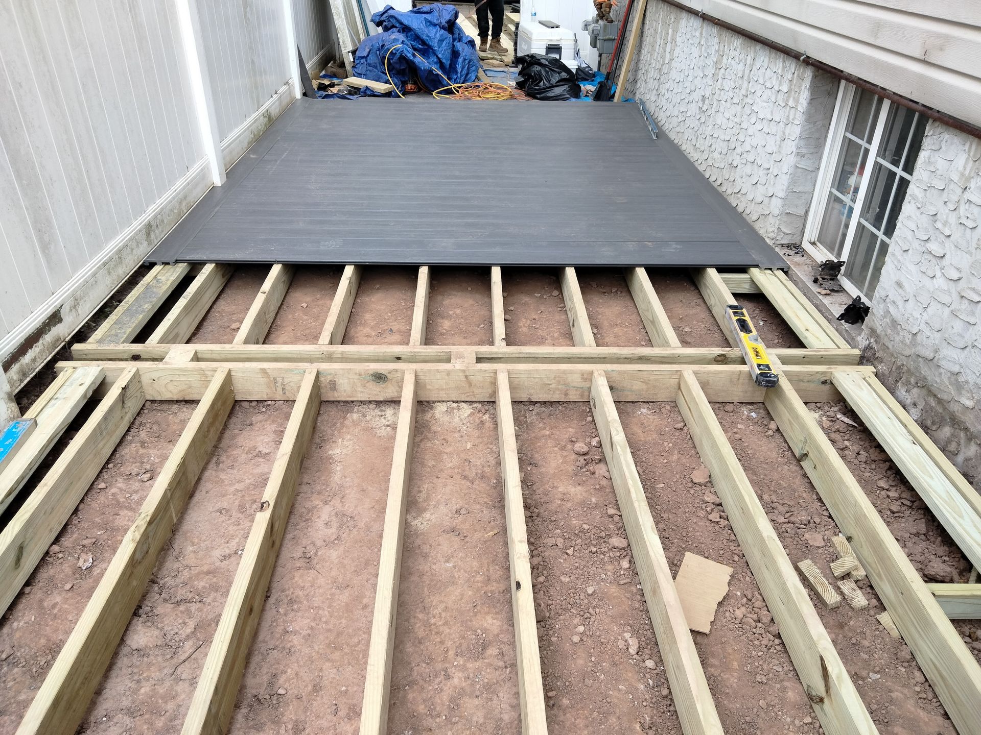 A deck under construction with gray composite planks installed over a wooden frame next to a house wall and white fence.