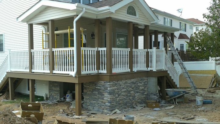A residential deck under construction featuring a gable roof, white railings, and a stone-veneer foundation.